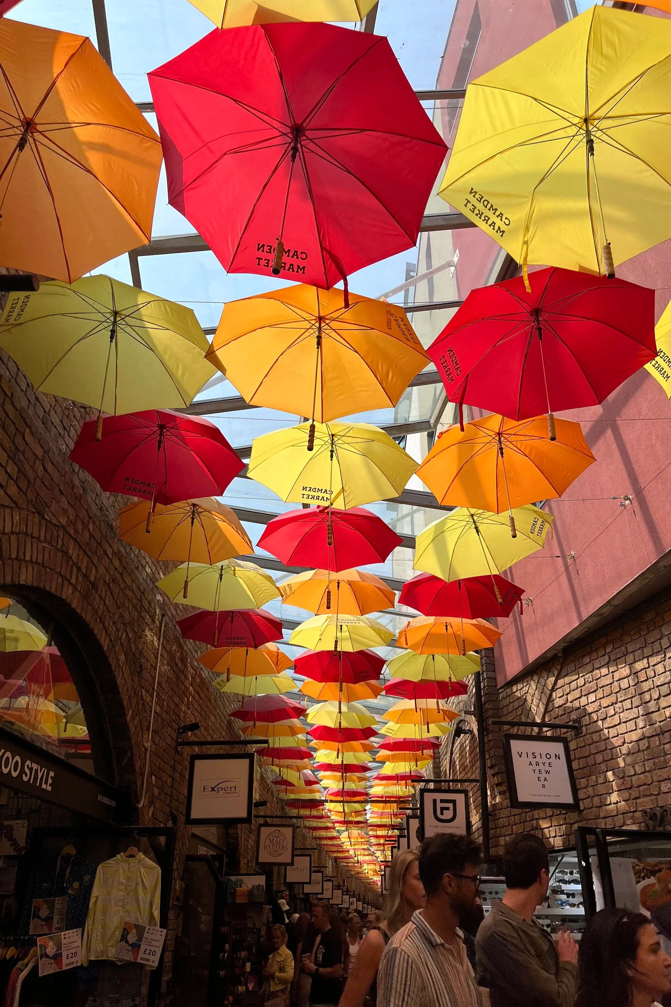 Tetto di ombrelli colorati (gialli, rossi, arancioni) sospesi sopra un mercato o un centro commerciale con persone che camminano sotto. Camden Market a Londra