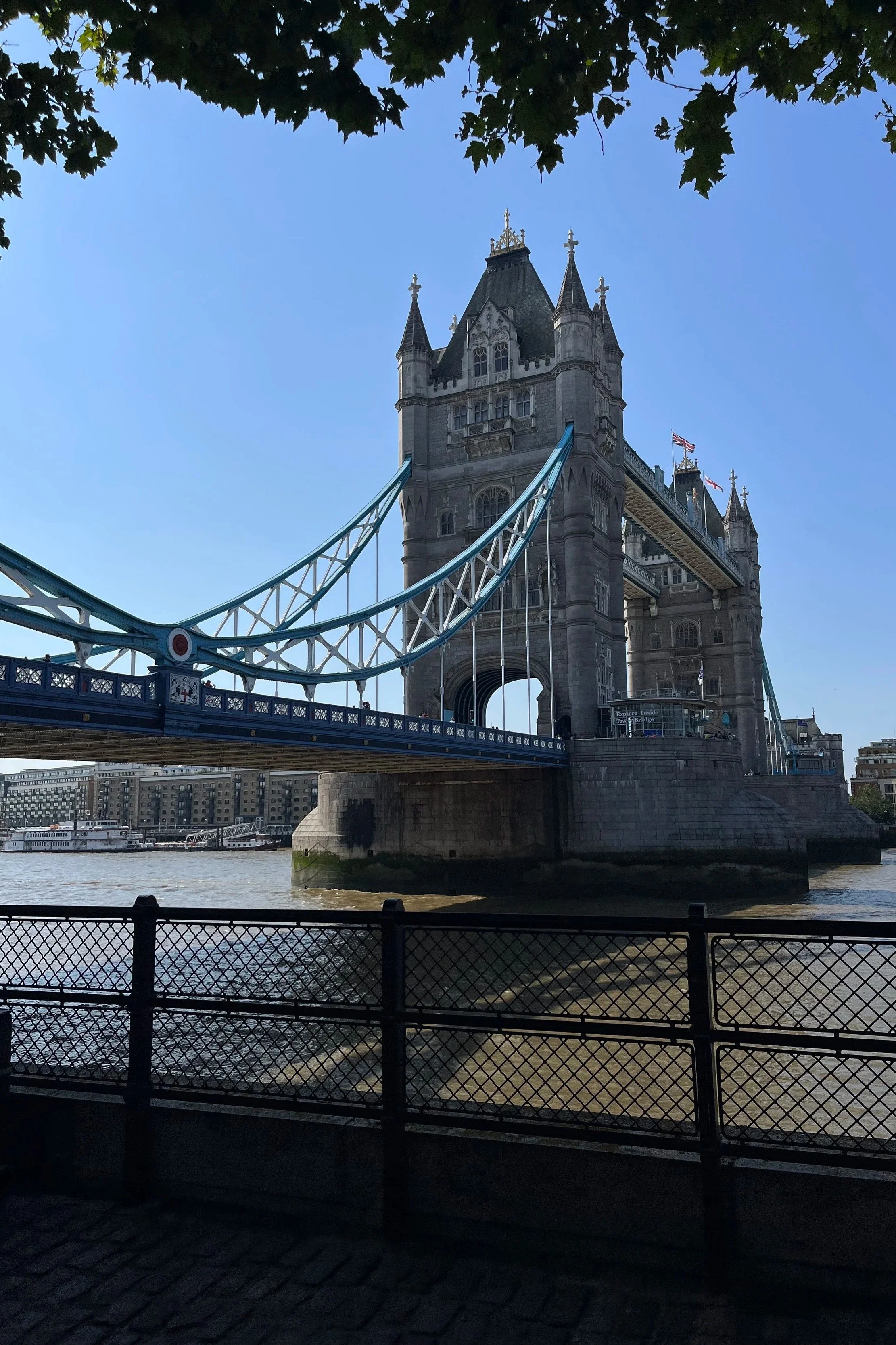 Il Tower Bridge di Londra visto dal lungotevere con il cielo azzurro e alcune foglie di alberi sopra.