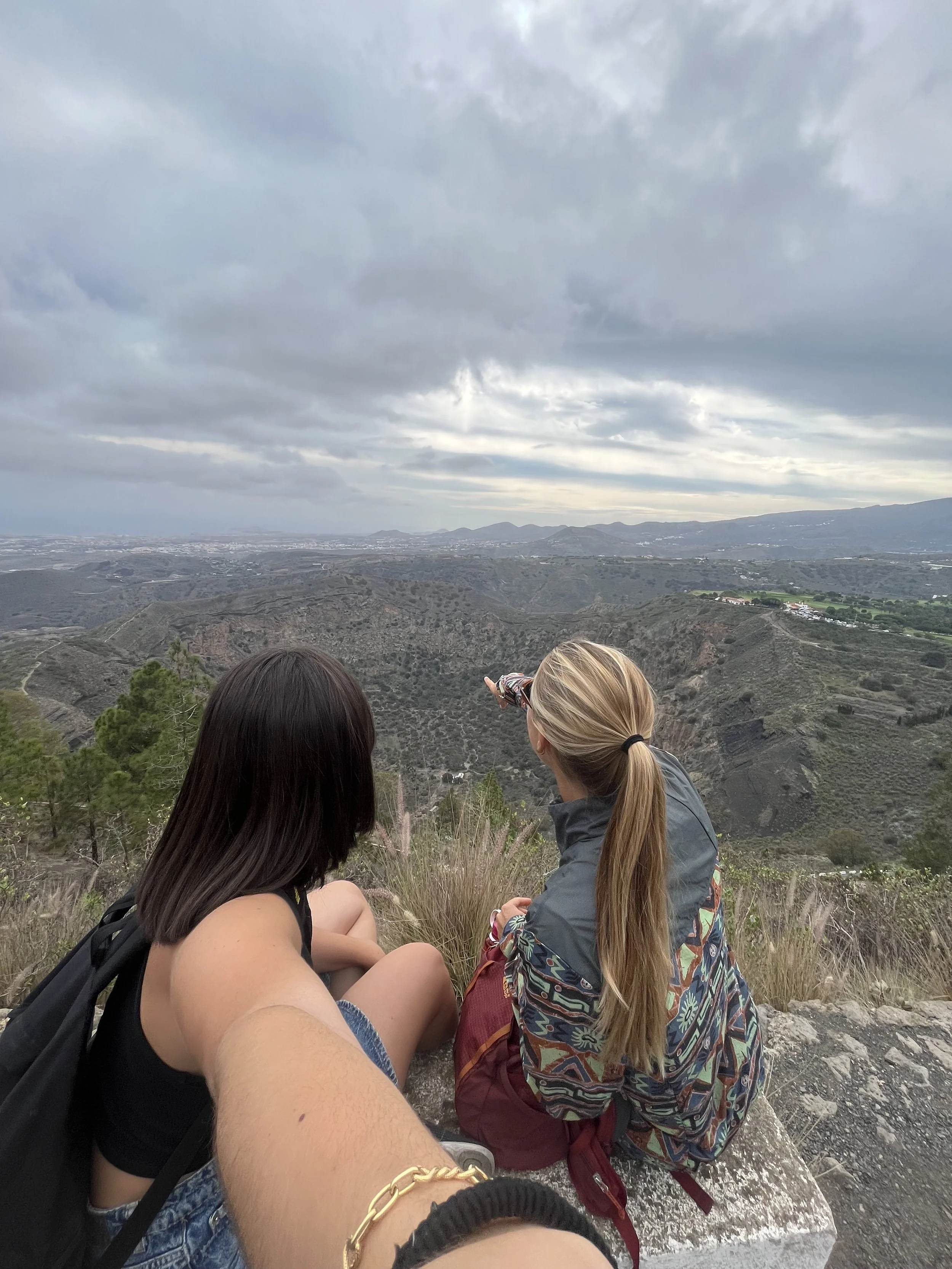 Due donne sedute su una roccia con vista su un paesaggio di colline e montagne sotto un cielo nuvoloso.