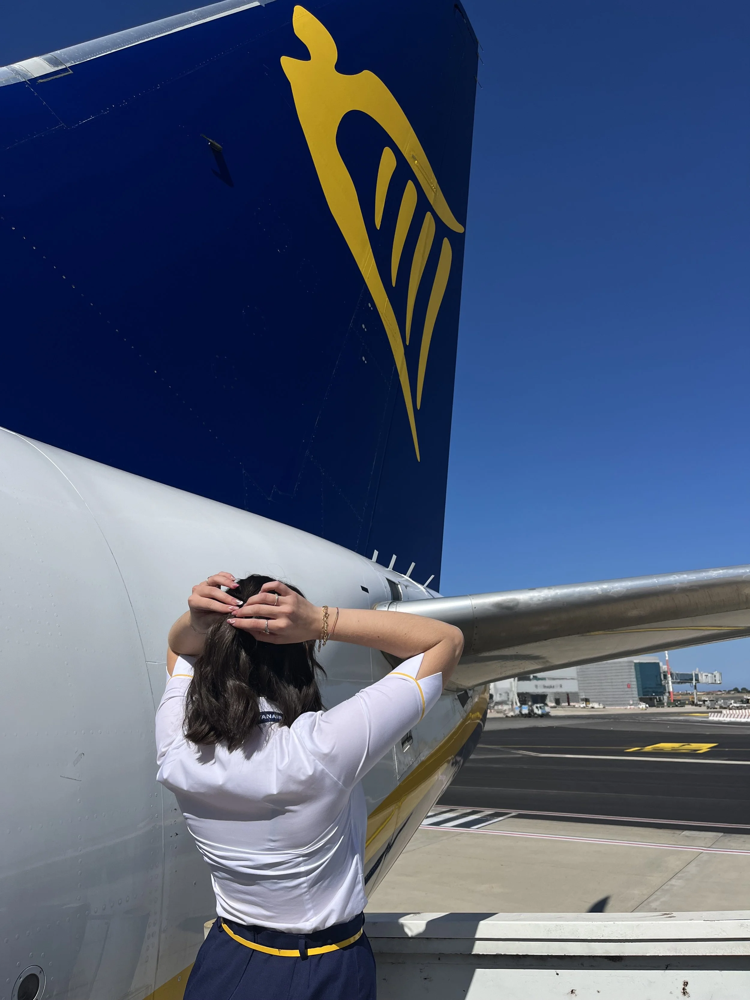 Donna in divisa di compagnie aerea, che si trova accanto a un aereo Ryanair in aeroporto, con le mani sulla testa, sotto un cielo blu.