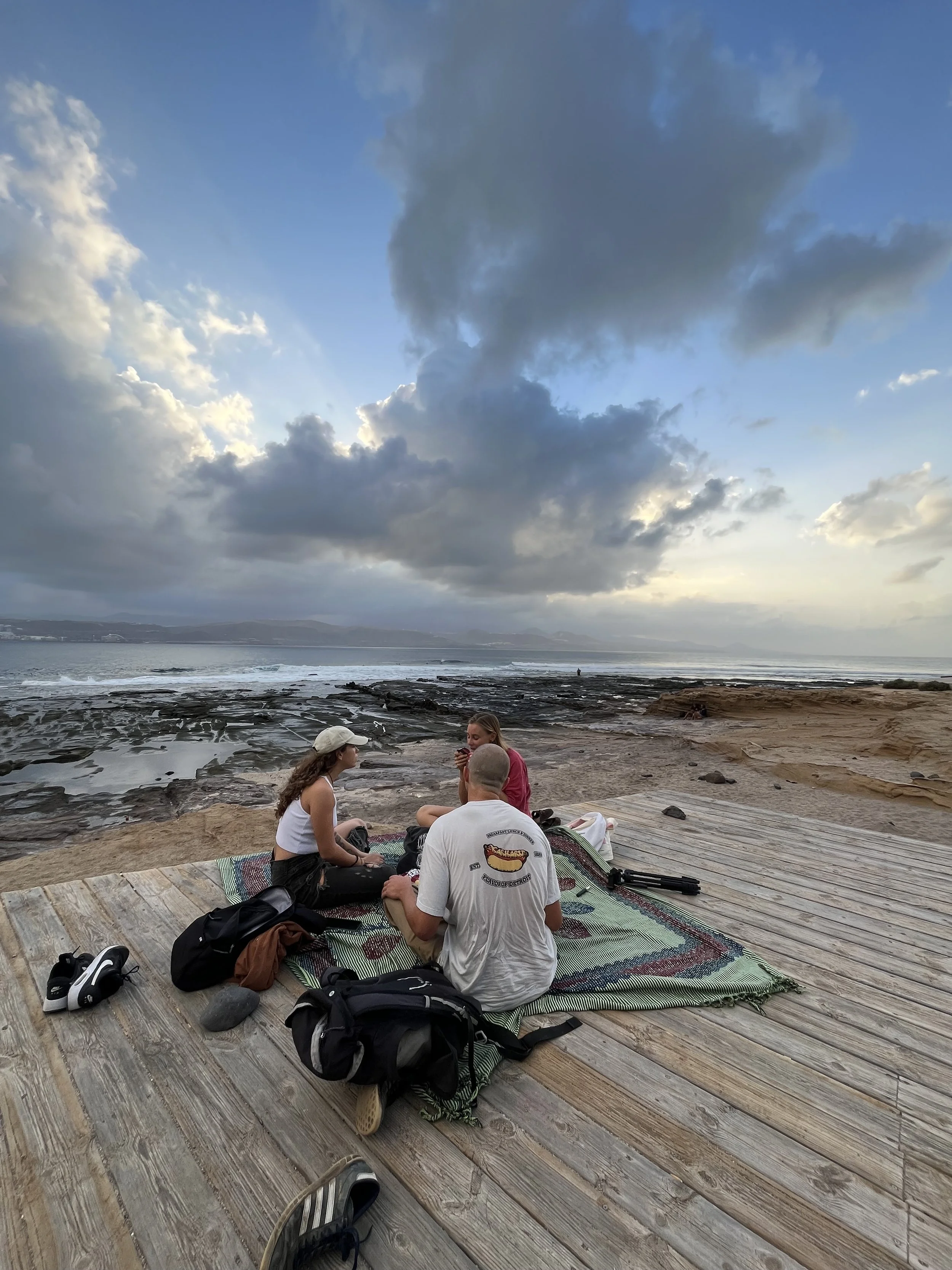 Tre persone sedute su un tappetino sulla banchina vicino alla spiaggia al tramonto, con cielo nuvoloso e mare in lontananza.
