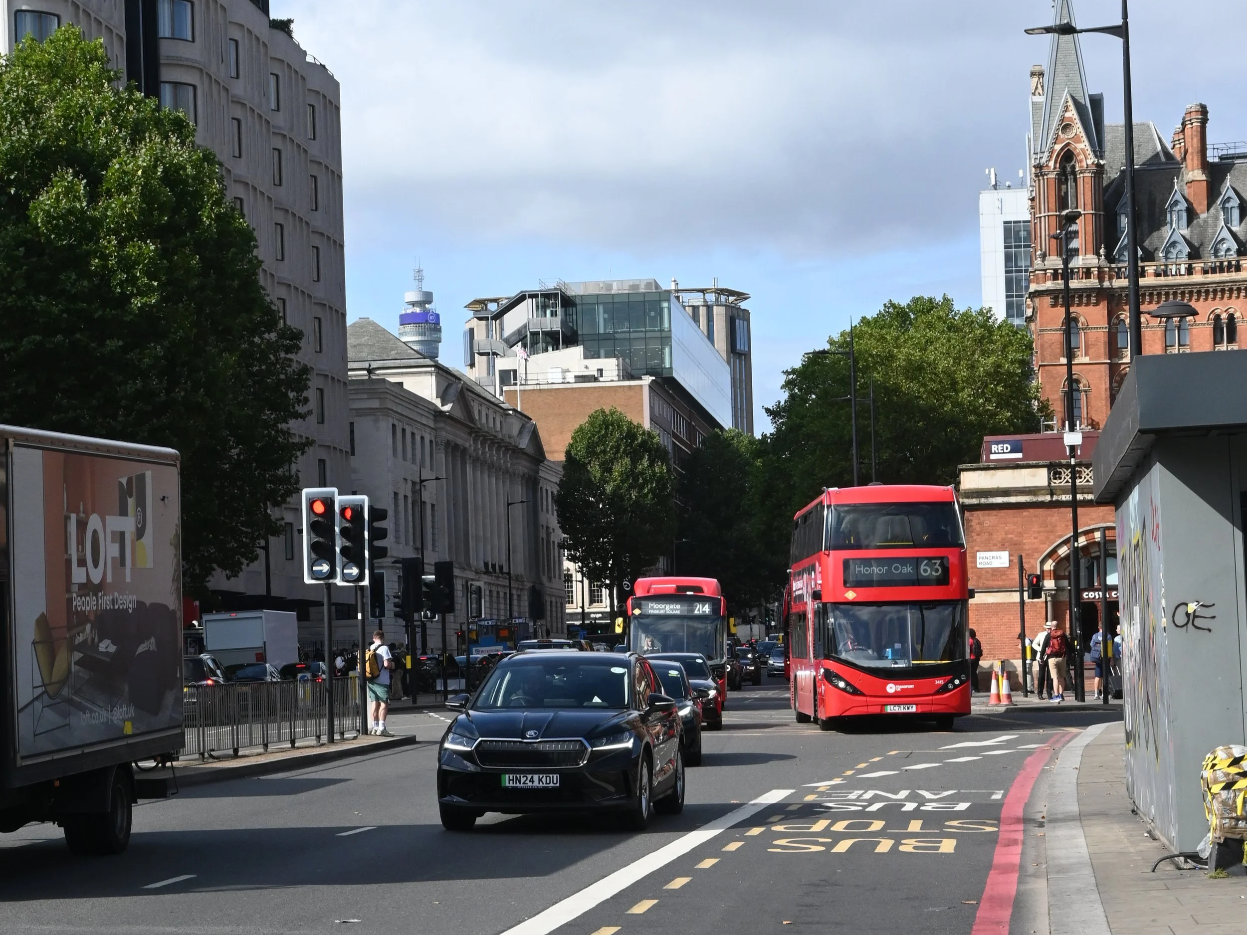 Vista di una strada urbana con autobus rossi, automobili, semafori e edifici storici e moderni, con alberi e cielo soleggiato, a Londra