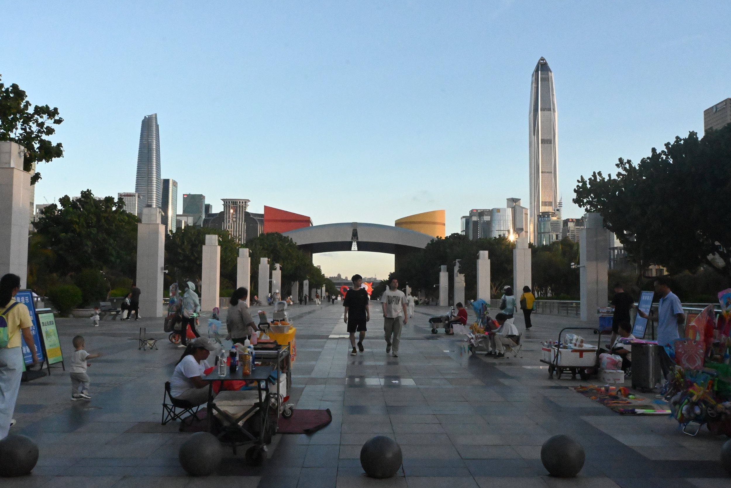 Una passeggiata nel parco con grattacieli moderni sullo sfondo, bancarelle e persone che passeggiano.