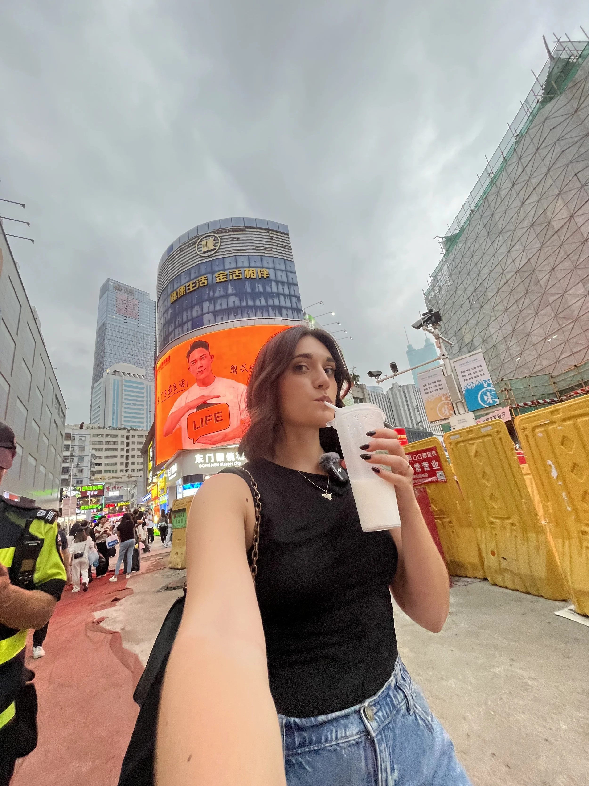 Una donna che prende un selfie in una zona urbana con edifici e schermi pubblicitari, mentre beve una bevanda ghiacciata con una cannuccia.