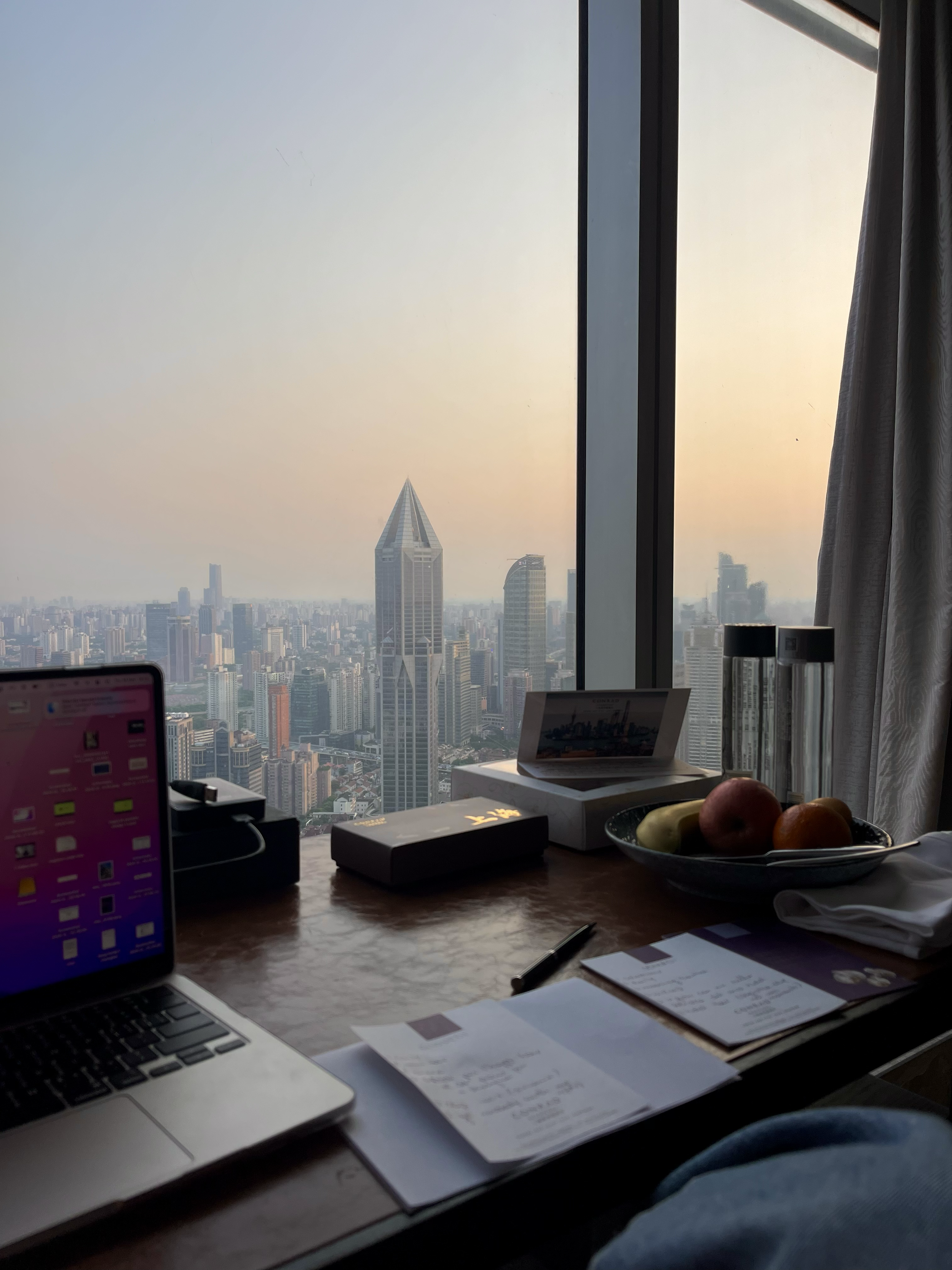 Vista di una città moderna attraverso una grande finestra con un tavolo vicino, che tiene un laptop, frutta, documenti, e due cilindri di vetro con liquido, con un cielo al tramonto. Ci troviamo al conrad hotel a shanghai.