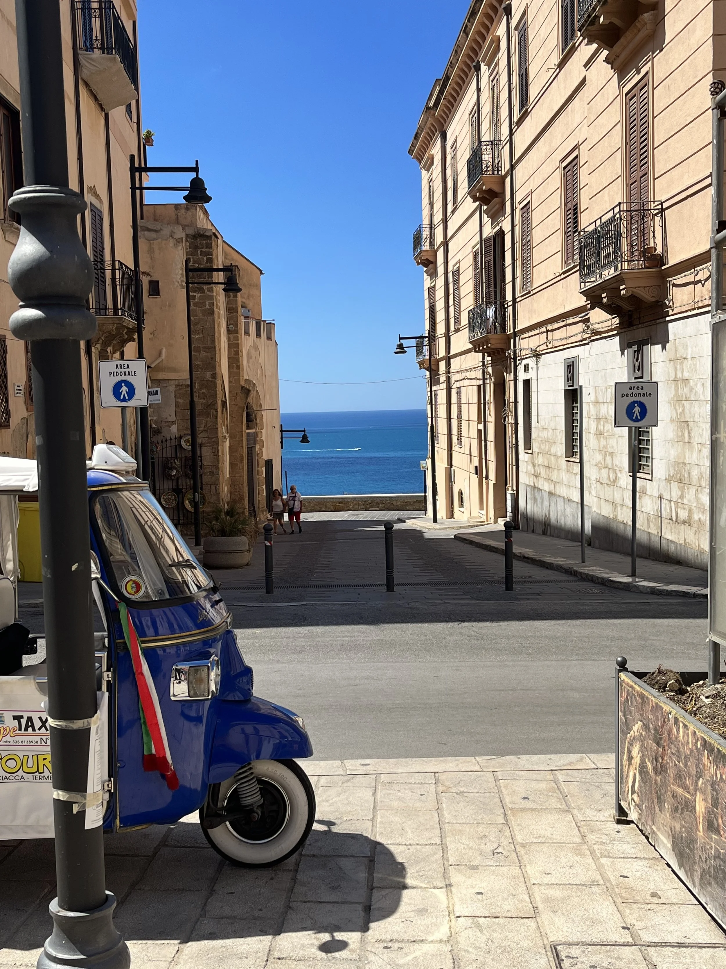 Vista di una strada stretta con edifici storici e balconi, che conduce al mare con il cielo blu sullo sfondo.