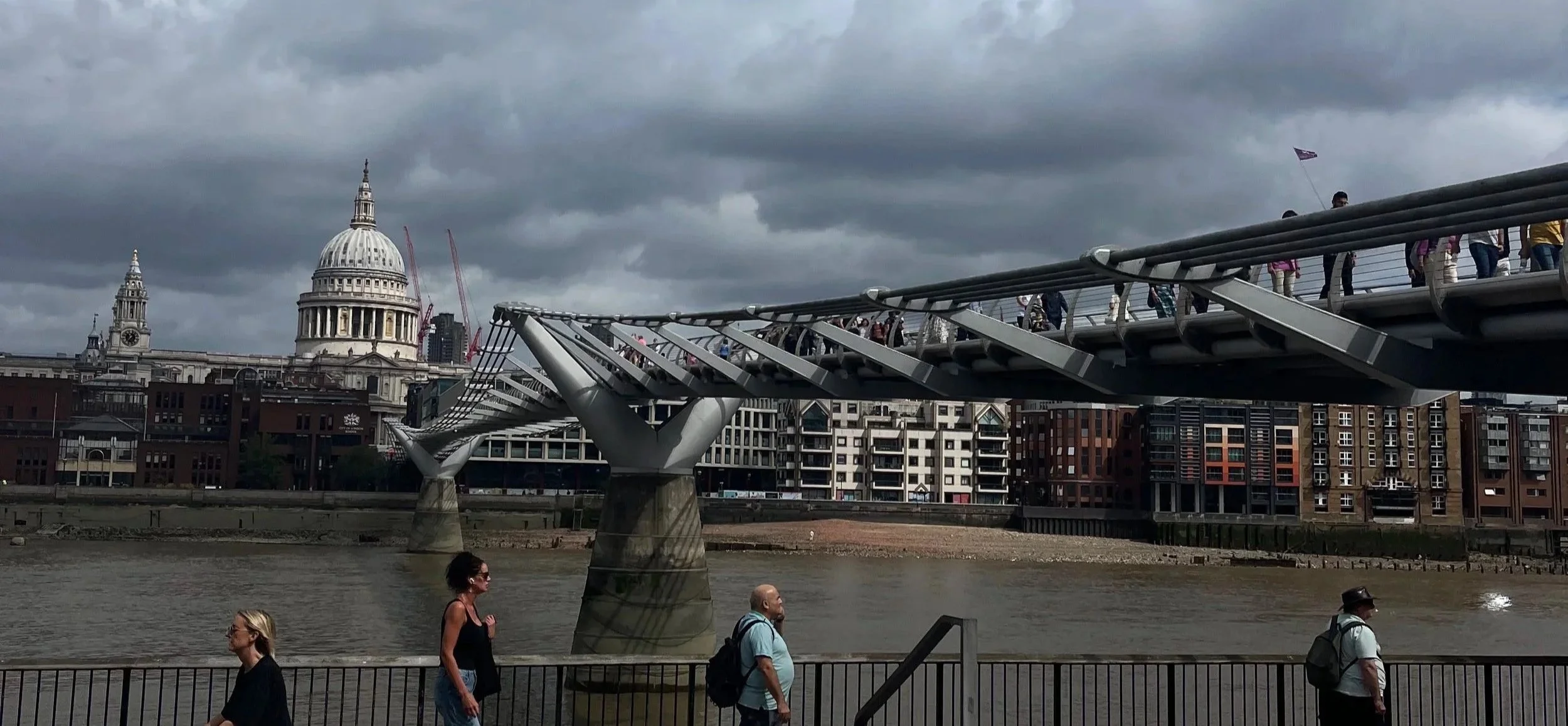 Veduta del Millennium Bridge sopra il fiume Tamigi con il municipio di Londra e la Cattedrale di St. Paul sullo sfondo, sotto un cielo nuvoloso.