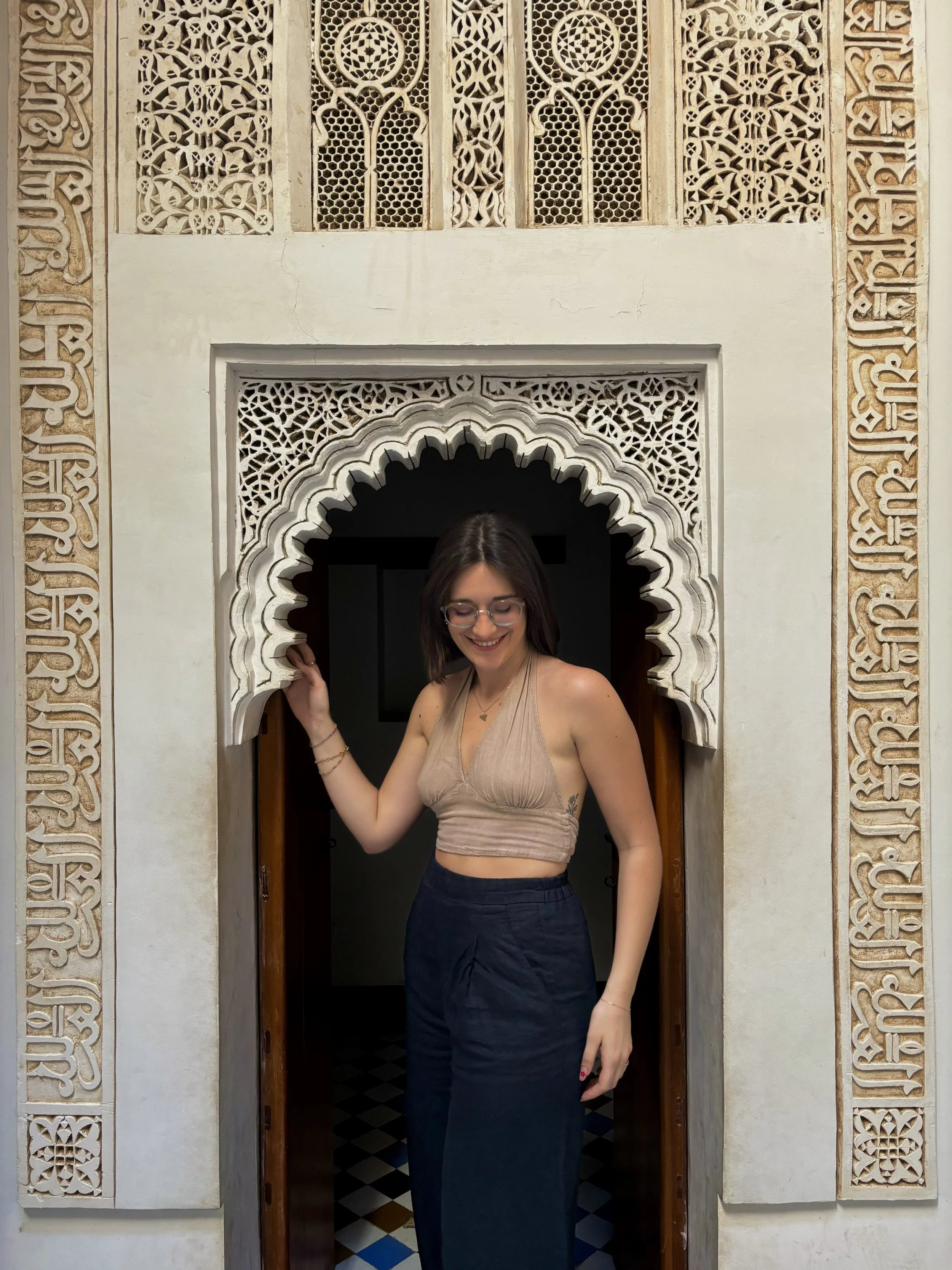 Una giovane donna con occhiali, capelli corti e vestiti in stile estivo si trova in una porta decorata in stile marocchino.