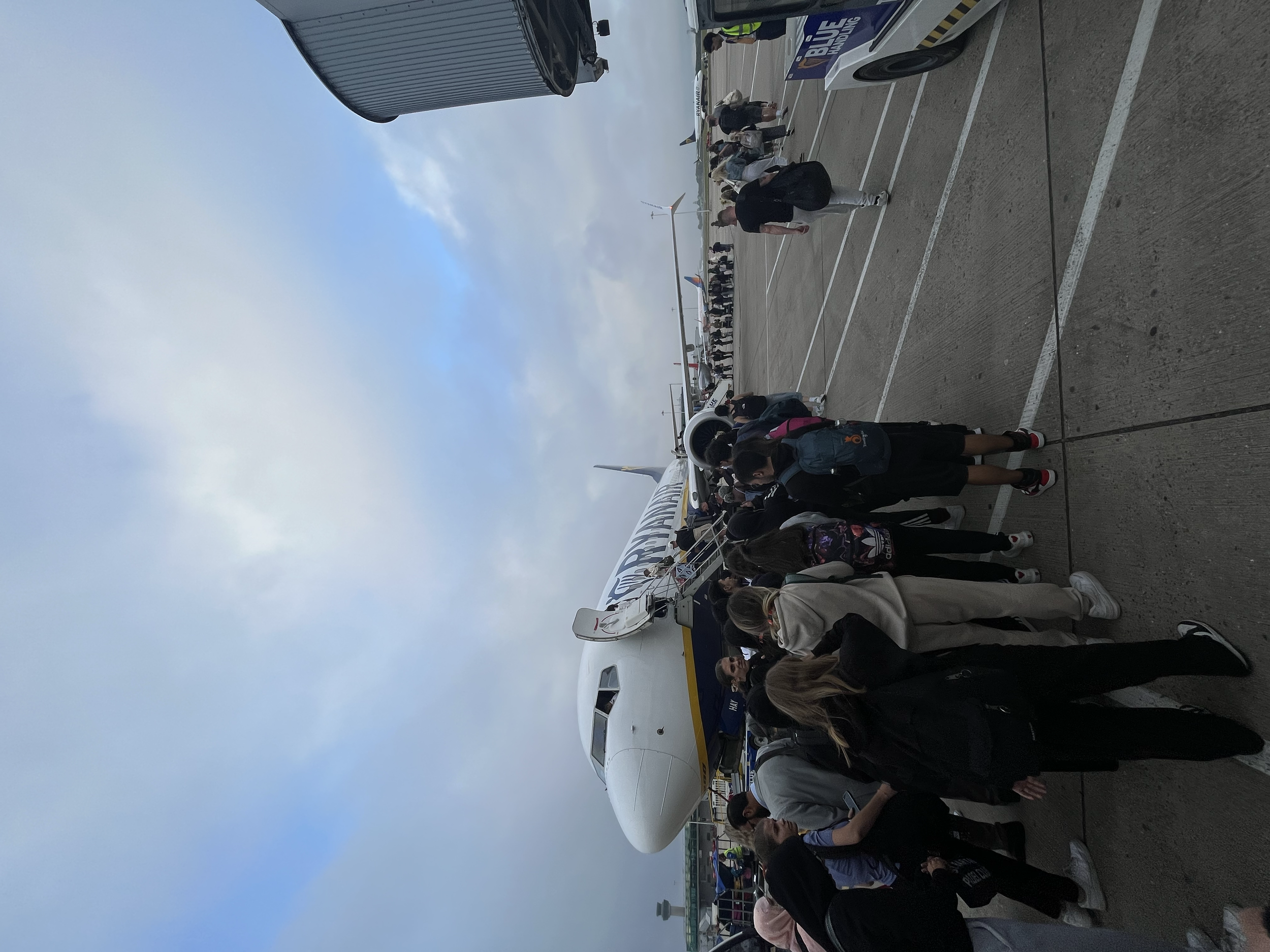 Gruppo di persone in fila vicino a un aereo Ryanair di linea sul molo, con cielo parzialmente nuvoloso.