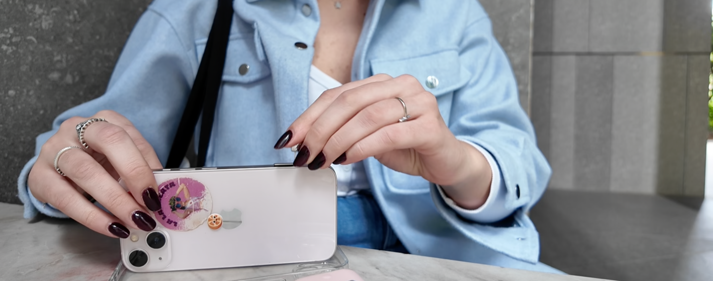 Una donna con un giubbotto di jeans chiaro sta usando uno smartphone decorato con adesivi, seduta a un tavolo con uno sfondo di pareti di cemento.