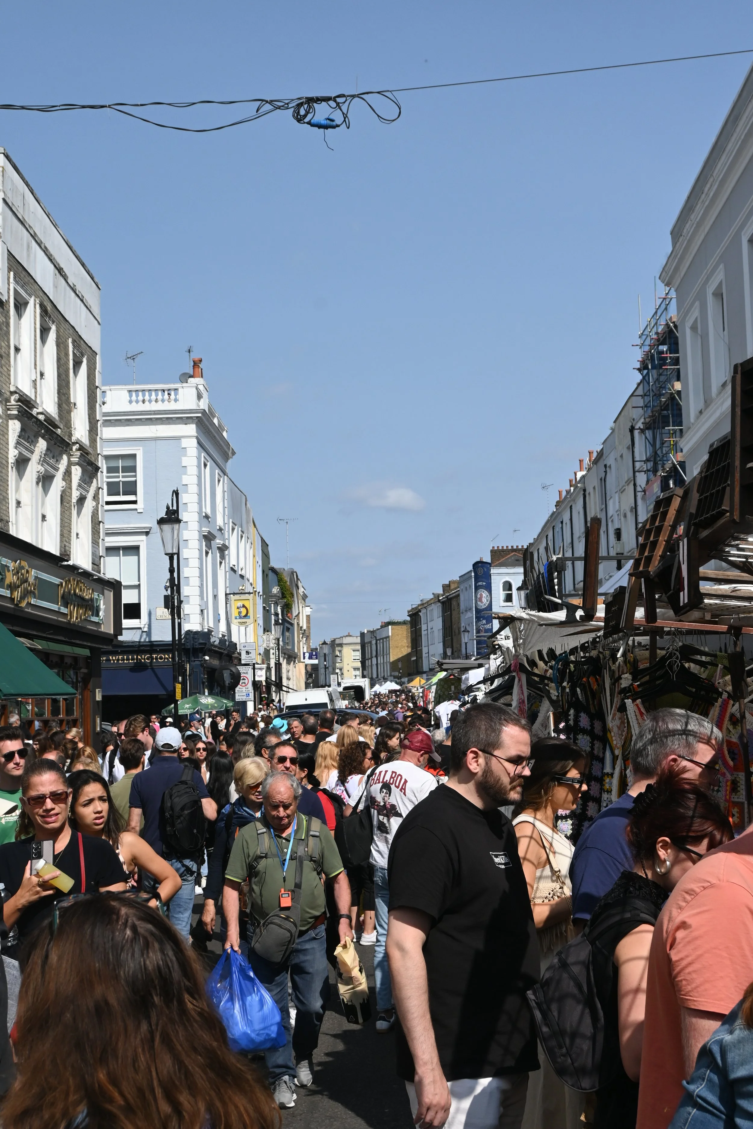 Via affollata di persone che passeggiano durante una fiera all'aperto, con negozi e bancarellequ verso il fondo. Edifici storici e segnaletica commerciale sono visibili lungo la strada, sotto un cielo blu.