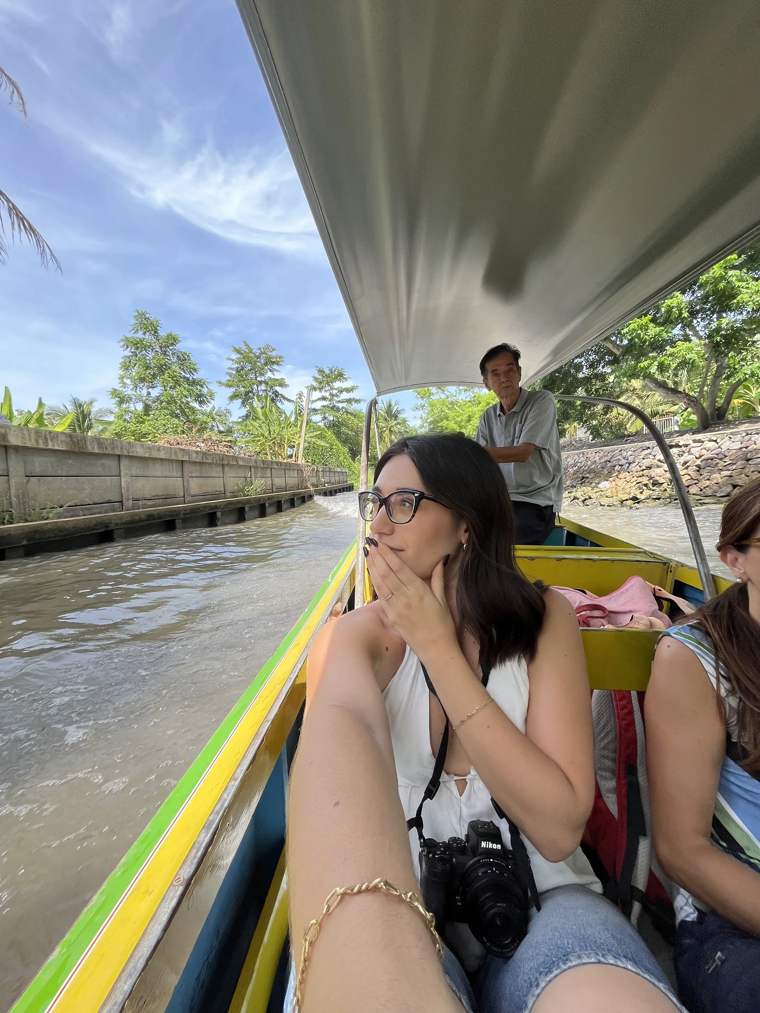 Persone sedute su una barca che attraversa un canale o fiume, con un uomo alla guida e un paesaggio verde e cielo azzurro sullo sfondo.