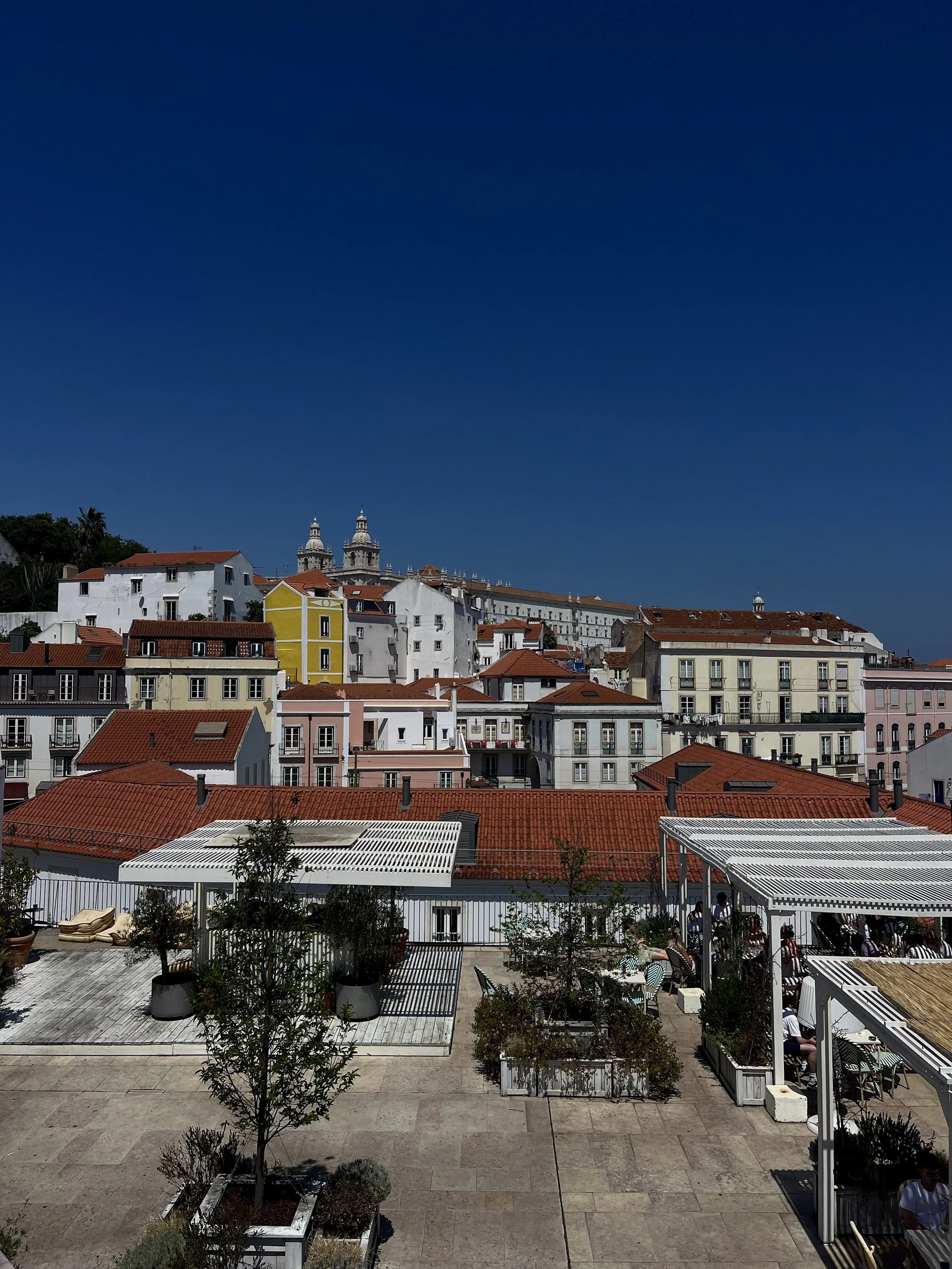 Veduta di un quartiere urbano con edifici colorati e tetti di terracotta, sotto un cielo sereno e blu, con una terrazza in primo piano con piante e sedie.