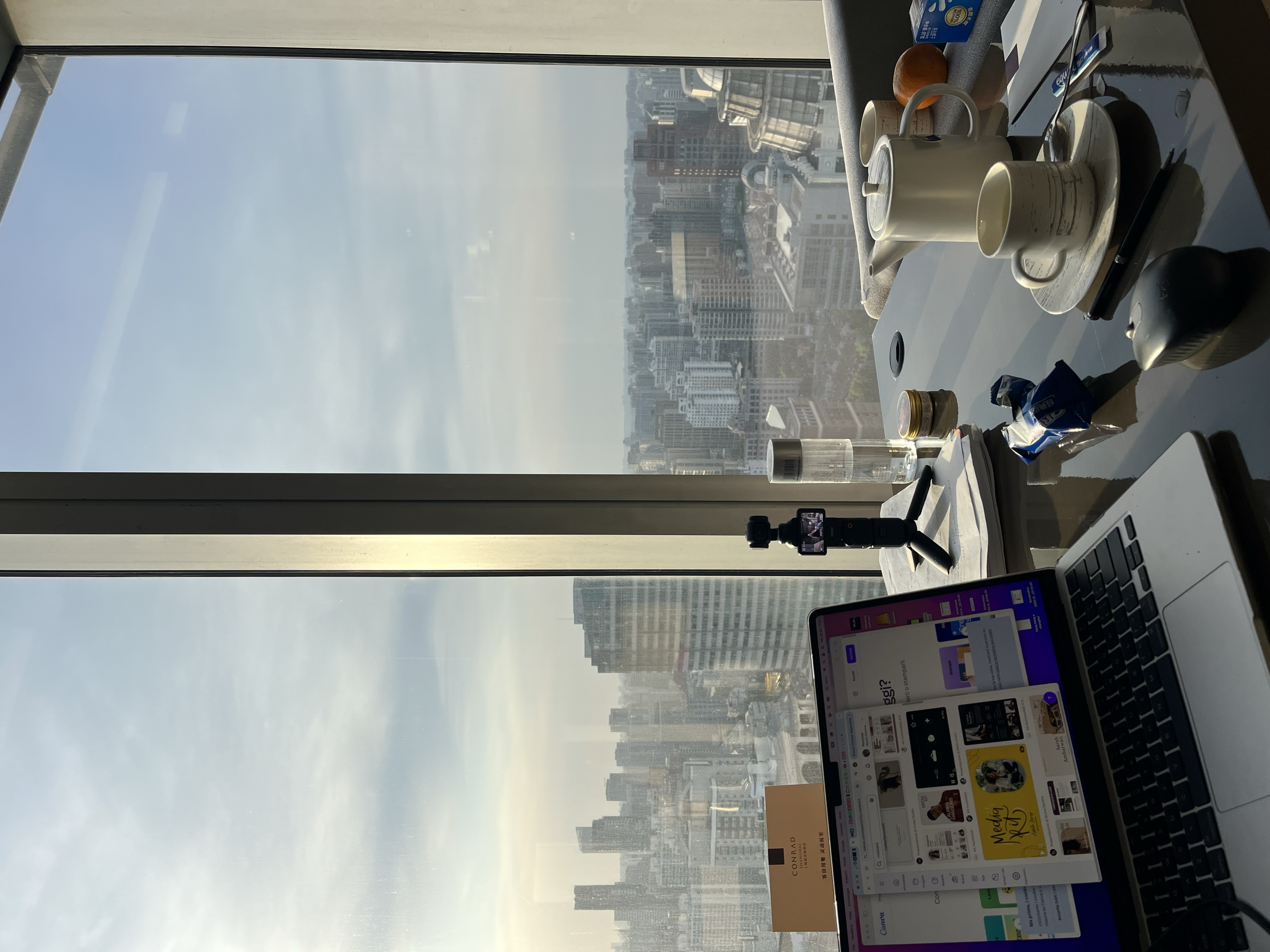 Scrivania con laptop, tazza, bottiglia d'acqua, snack, e gadget tecnologici, con vista sulla città attraverso una finestra alta. ci troviamo al conrad hotel a shanghai.