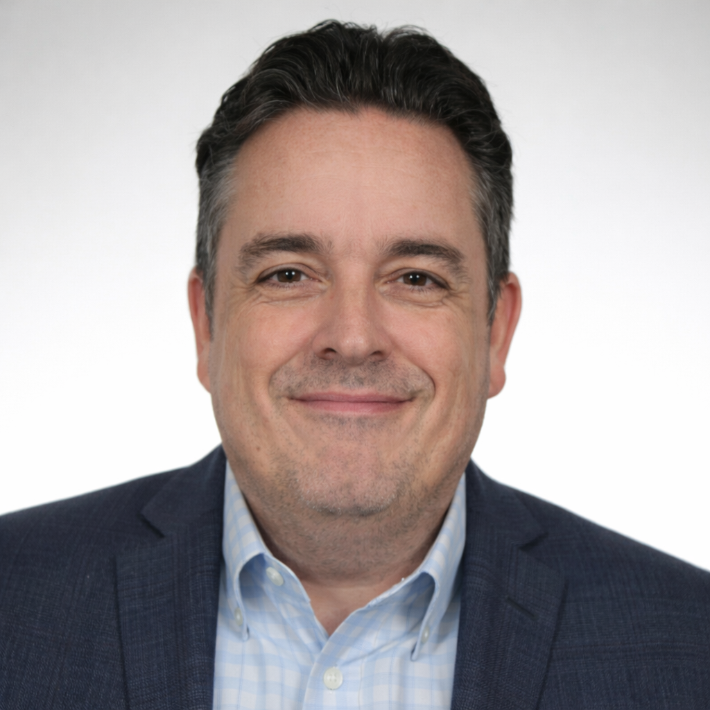 Headshot of a middle-aged man with dark hair, wearing a checkered dress shirt and dark blazer, smiling against a plain white background.