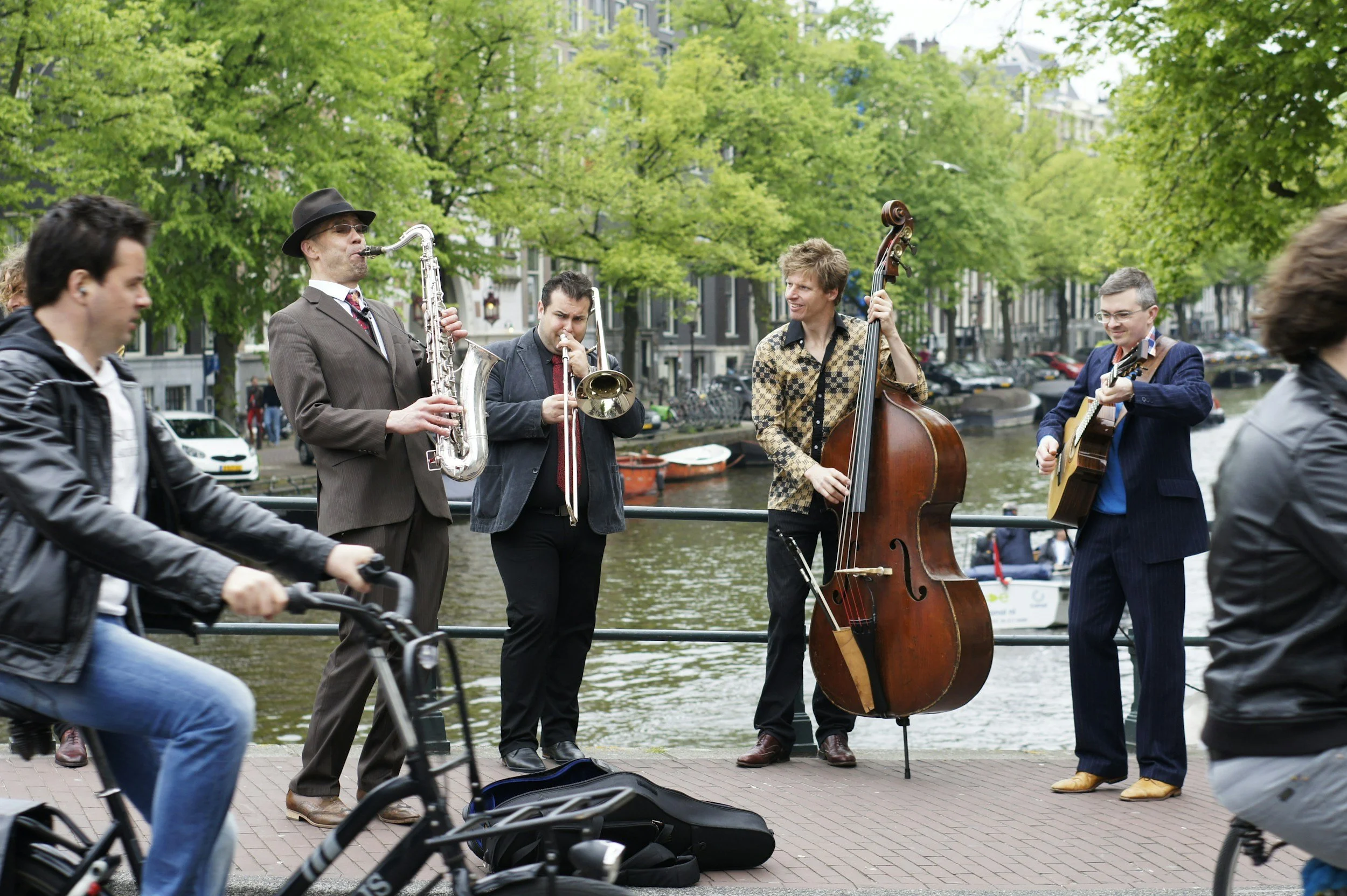 A street musician band playing jazz music by a canal in a city park, with pedestrians and cyclists passing by, green trees in the background.