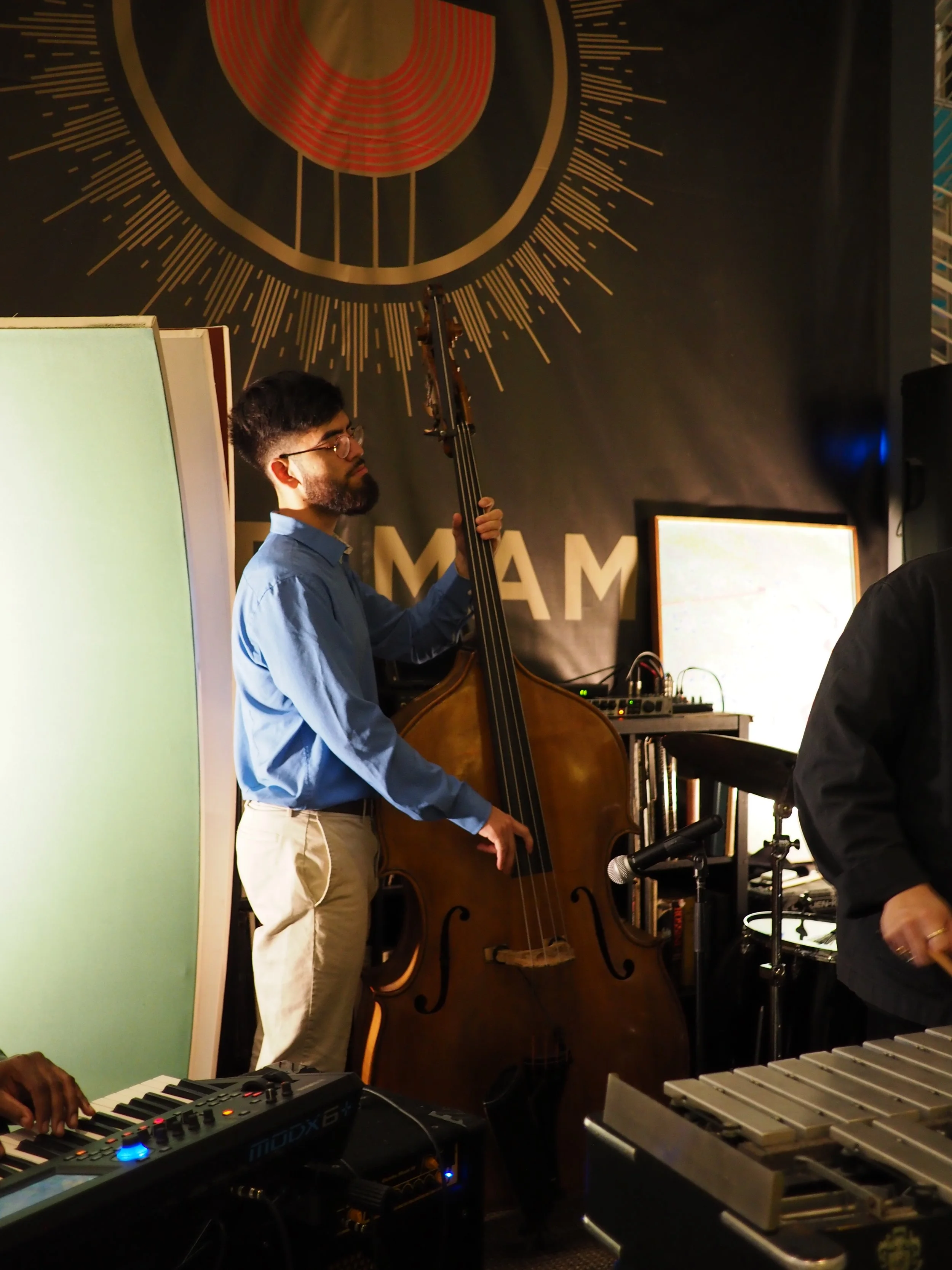 A man playing a double bass during a musical performance, with electronic keyboards and other musical equipment around him, indoors with a dark background and decorative wall art.