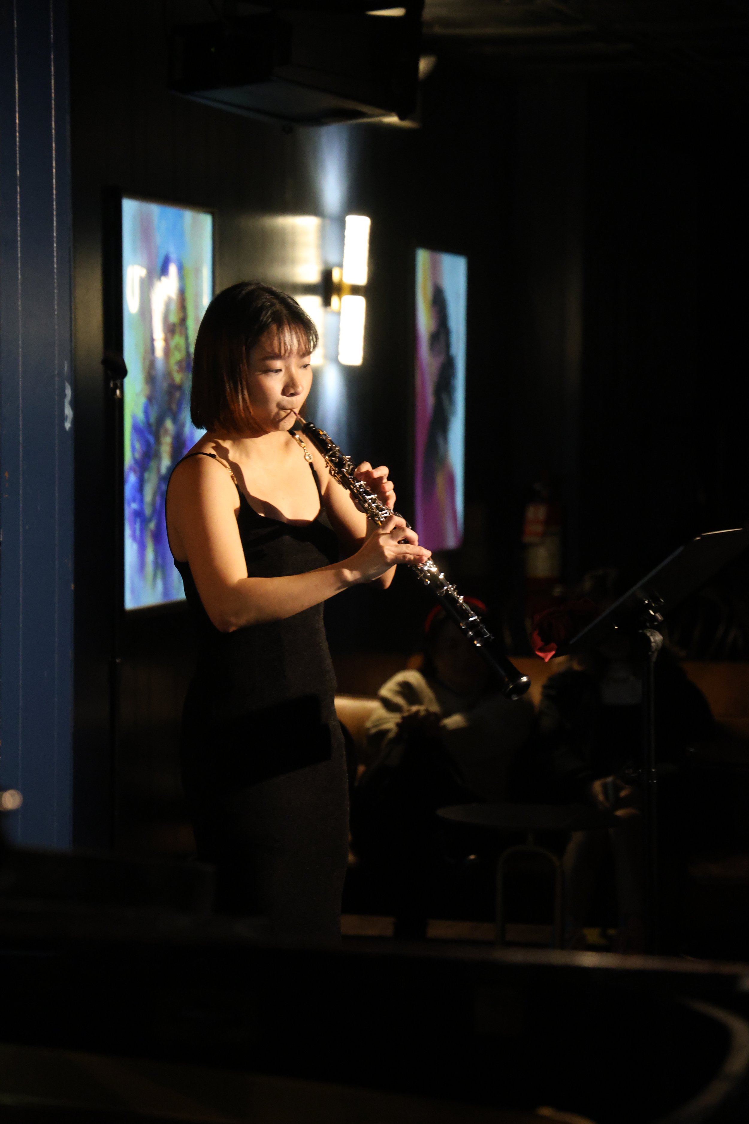 A woman in a black dress is playing an oboe on stage with dim lighting and colorful artwork in the background.