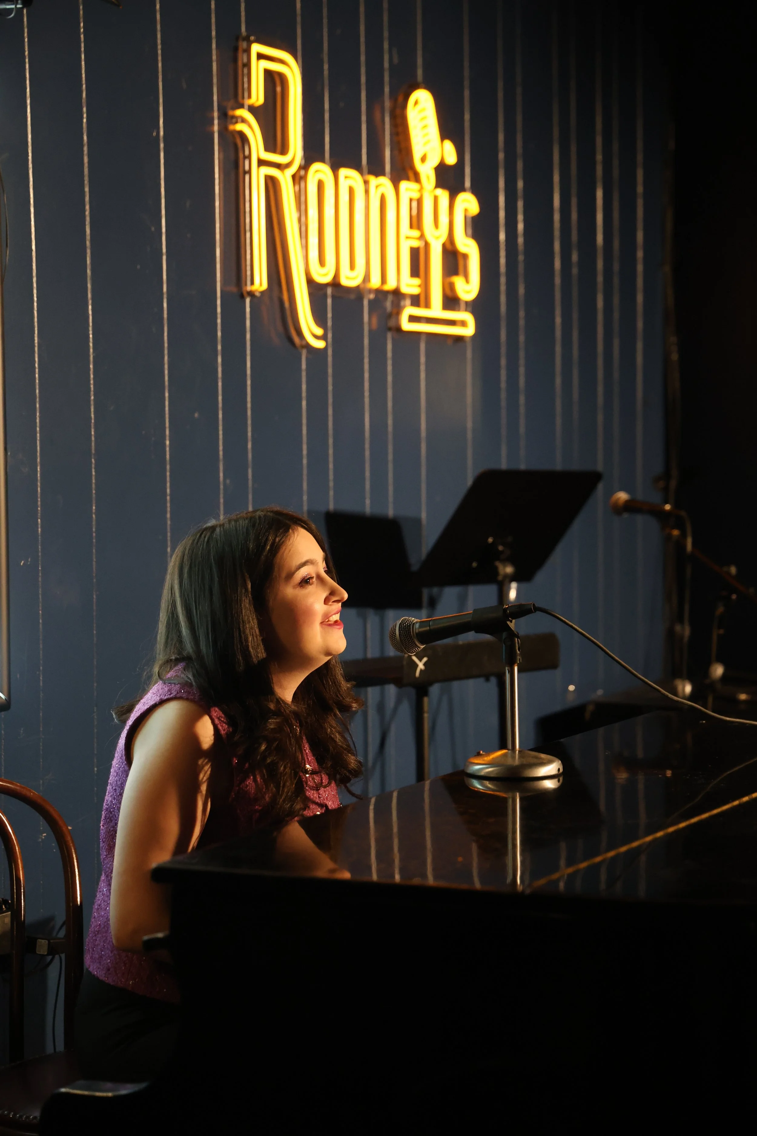 A woman with dark hair singing or speaking into a microphone at a piano in a dimly lit jazz club or bar, Rodeney's.
