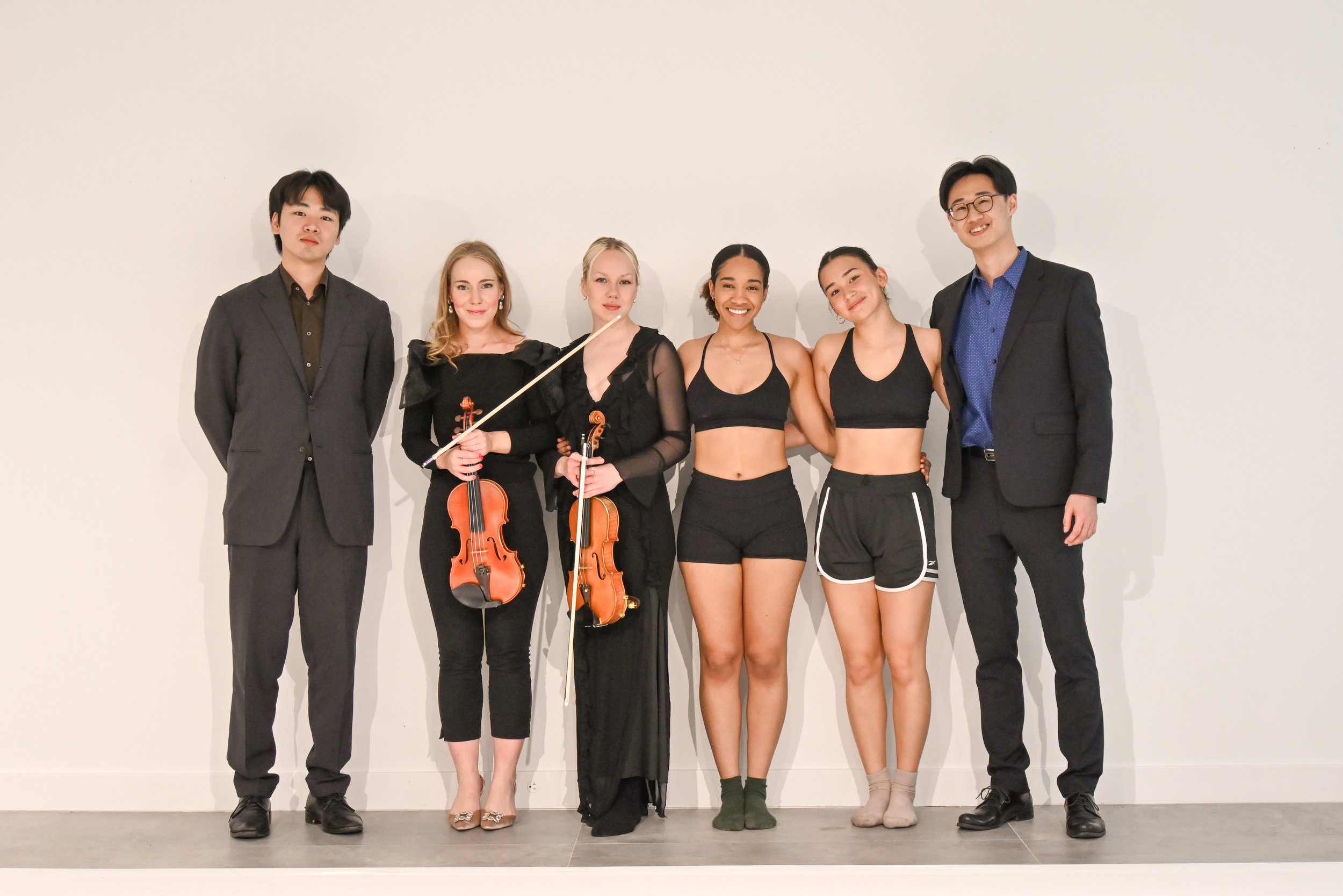 Group of five young people, three men and four women, standing against a plain white wall. Two women in the middle hold violins, and the others are dressed in casual and formal attire, smiling at the camera.