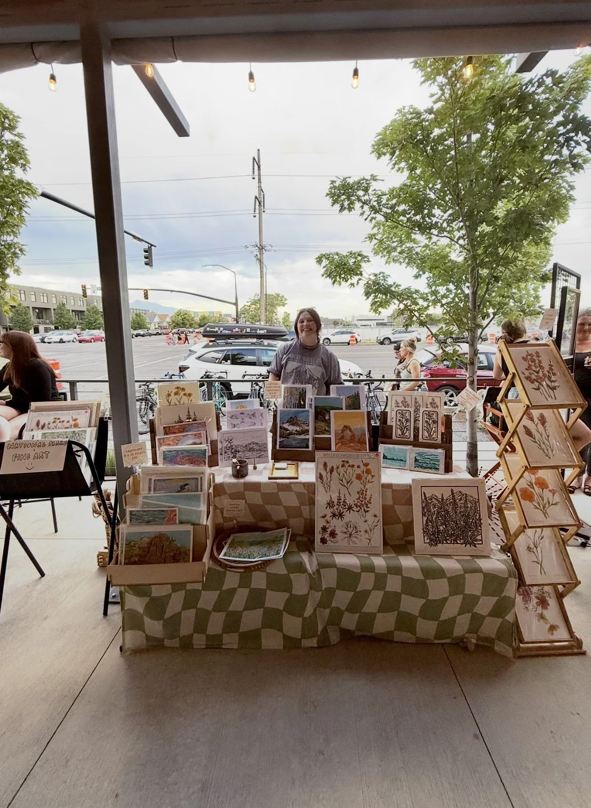 An outdoor art fair booth displaying botanical and landscape prints, with a smiling woman standing behind the table, a tree, and a parking lot with cars and people in the background.