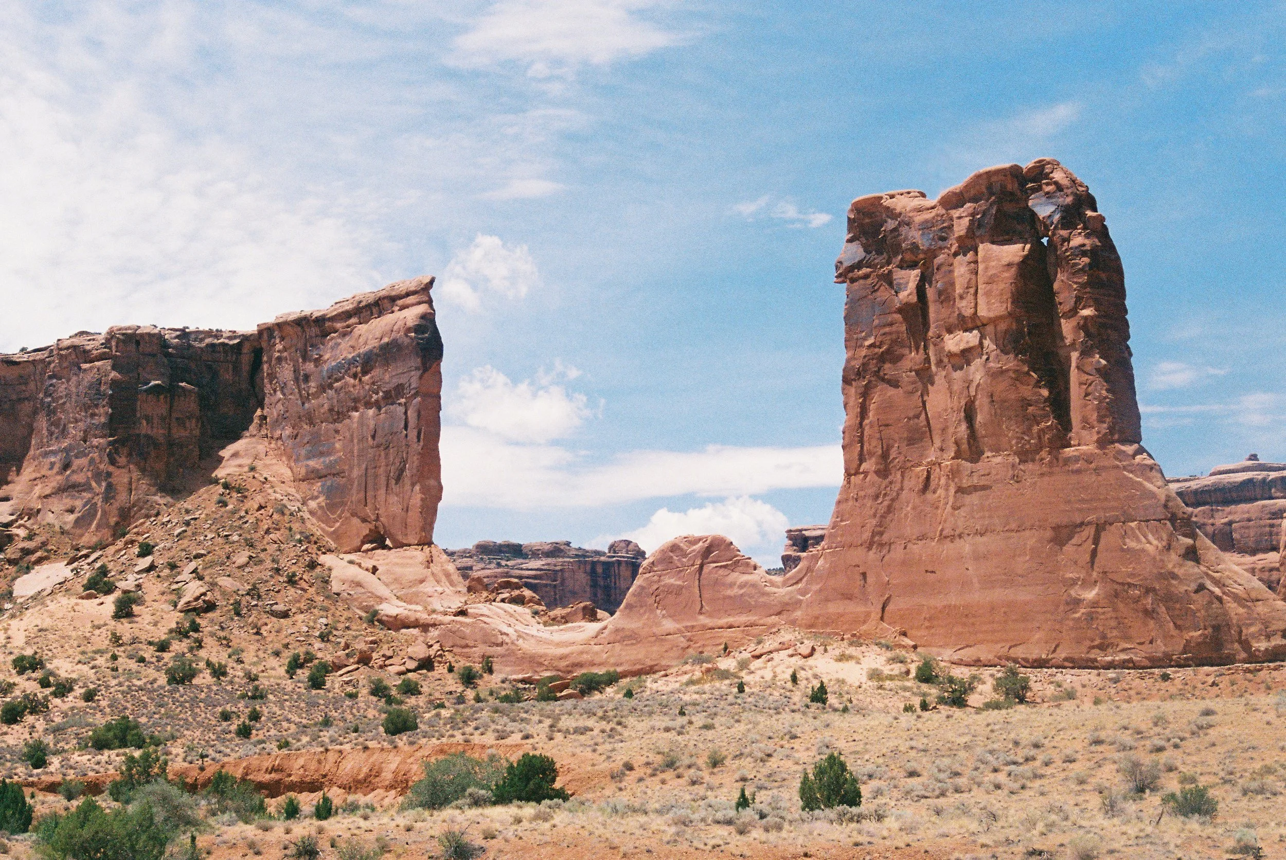 Arid desert landscape with prominent red rock formations and a partly cloudy blue sky.