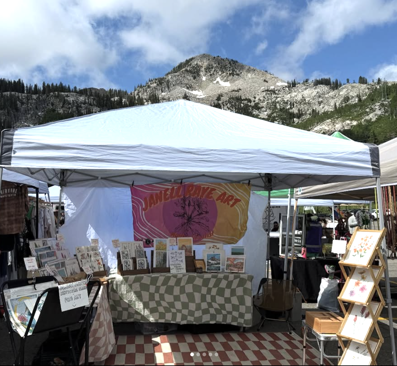 Outdoor craft vendor tent at a market with mountains and blue sky in the background, displaying artwork and crafts.