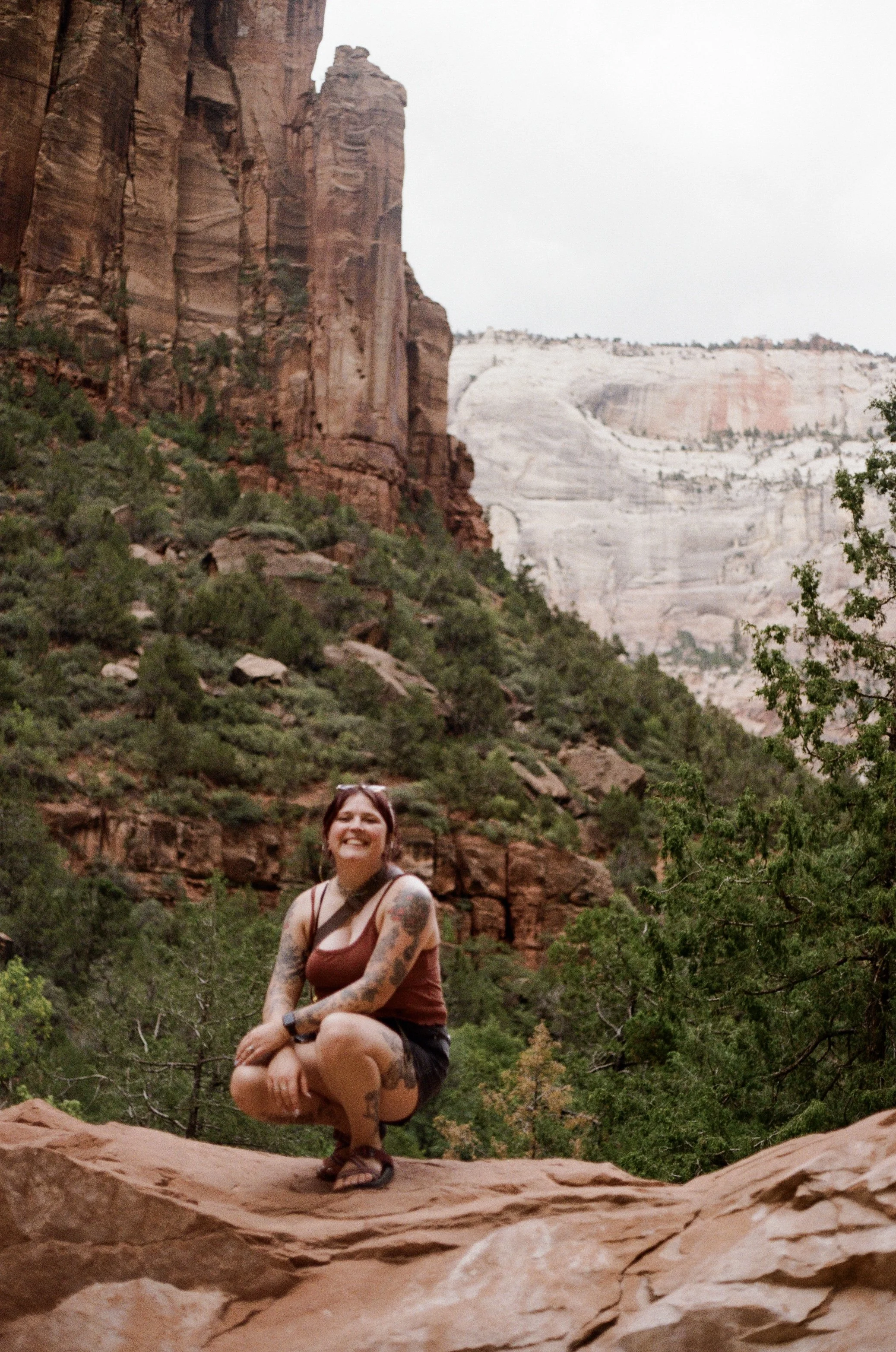 A woman with tattoos on her arms and legs, wearing a tank top and shorts, squatting on a rock in a canyon landscape with tall red and white cliffs and green vegetation.