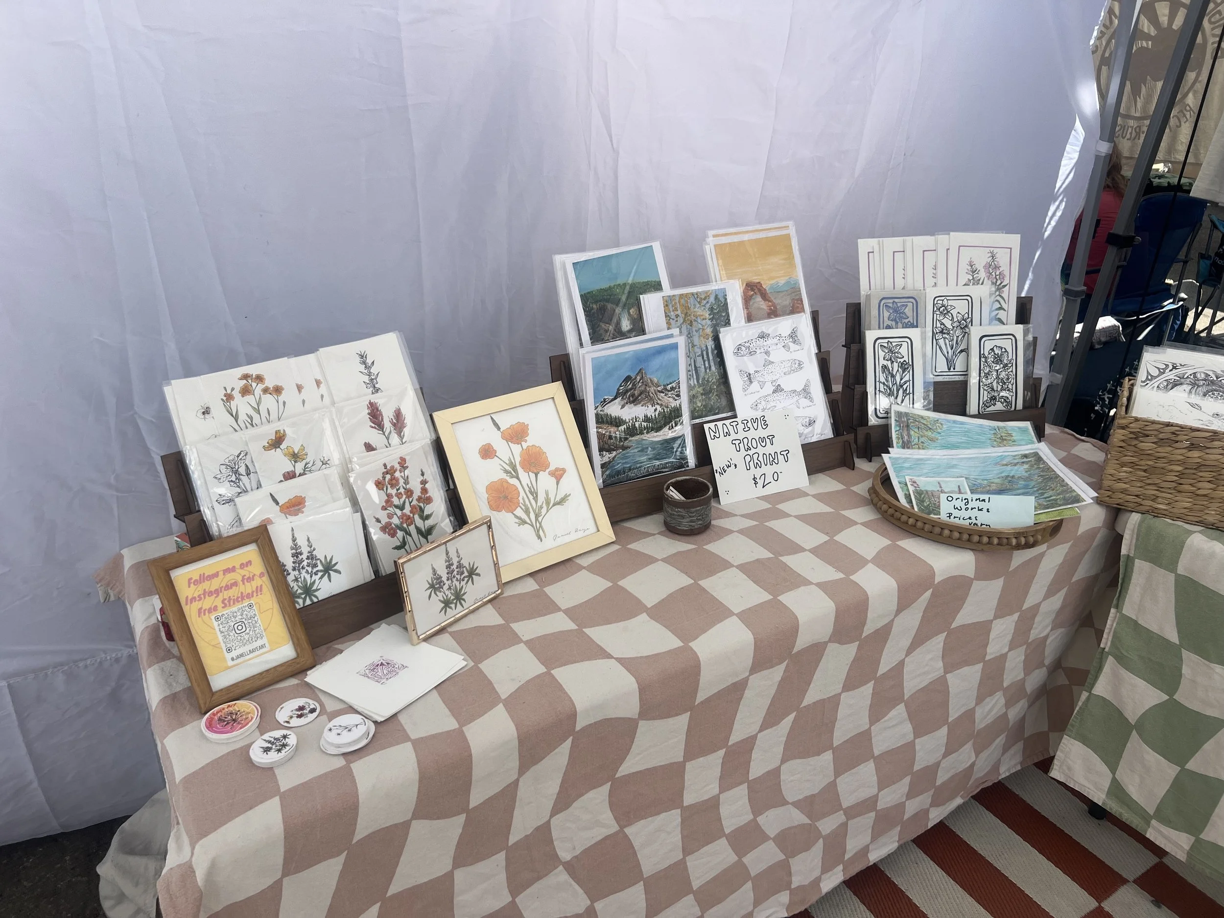 A display table at an outdoor market, featuring framed art prints of wildflowers and landscapes, small handmade crafts, and a sign advertising native trout art for $20, with a QR code for Instagram.