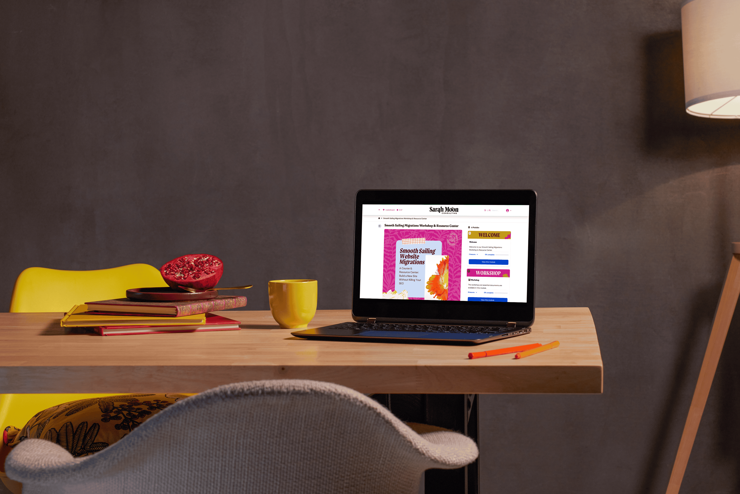 A workspace with a wooden desk, yellow chair, and a gray wall in the background. On the desk, there is a laptop displaying a webpage, a yellow mug, a pomegranate cut in half, and a stack of colorful notebooks. There are two orange pens on the desk, and a cushioned chair with a patterned fabric is partially visible.