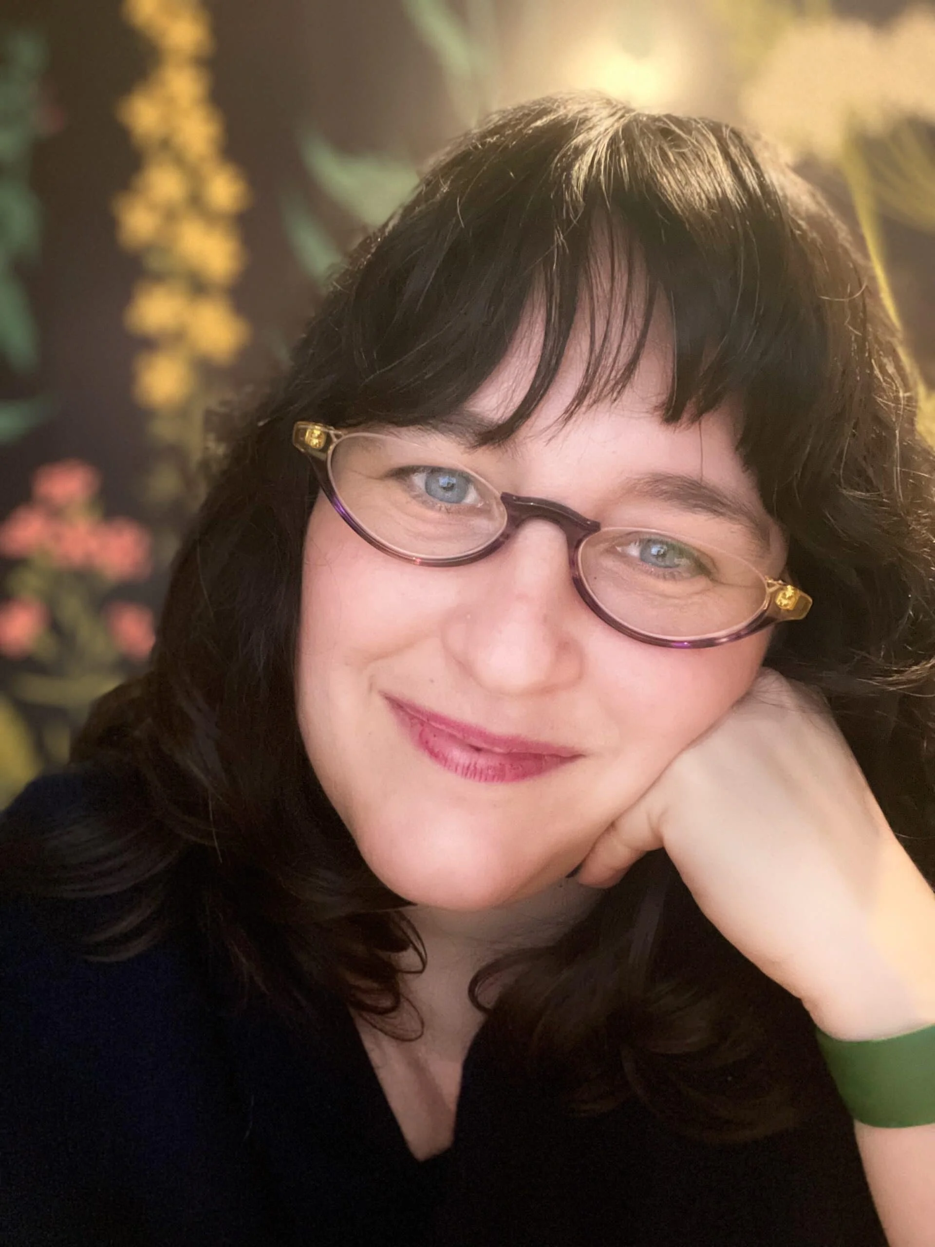 Close-up of a woman with dark curly hair wearing glasses, resting her chin on her hand, smiling at the camera, with a floral backdrop.