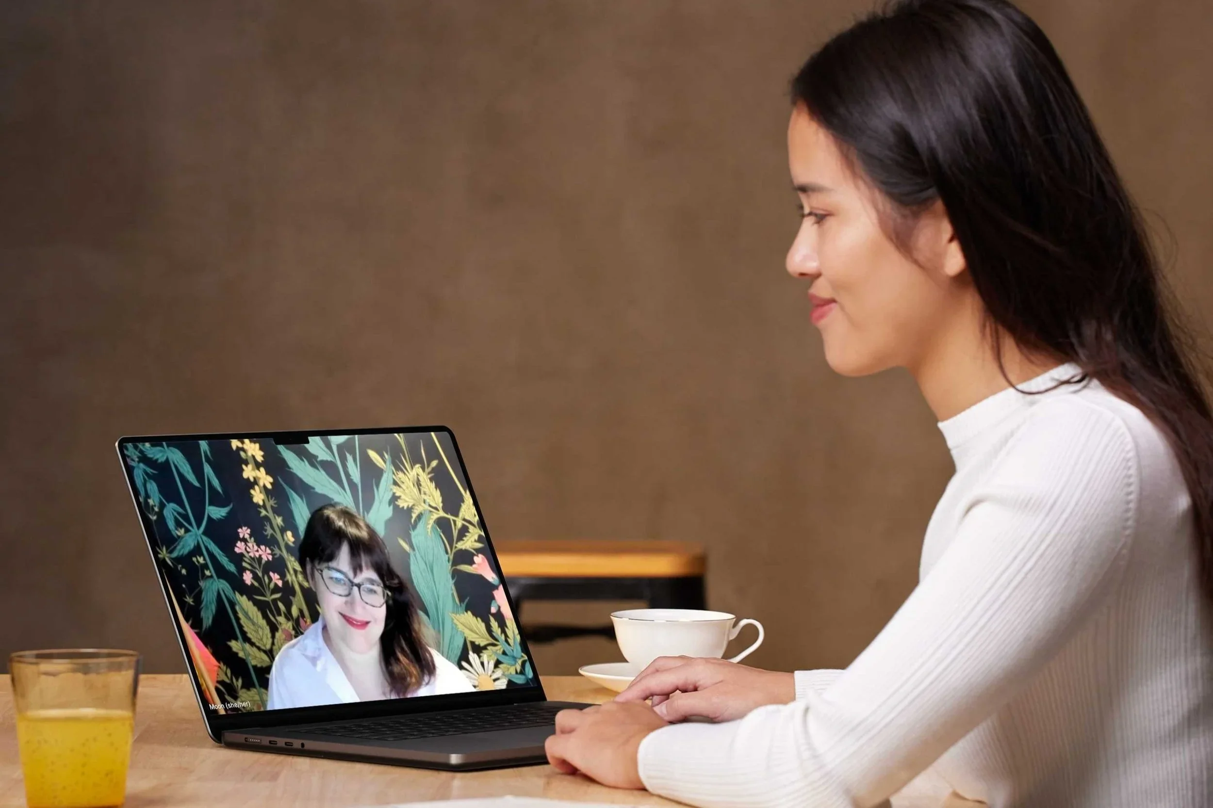 A woman with dark hair sitting at a table, using a laptop for a video call with another woman who has glasses and wavy hair, against a colorful botanical wallpaper background.