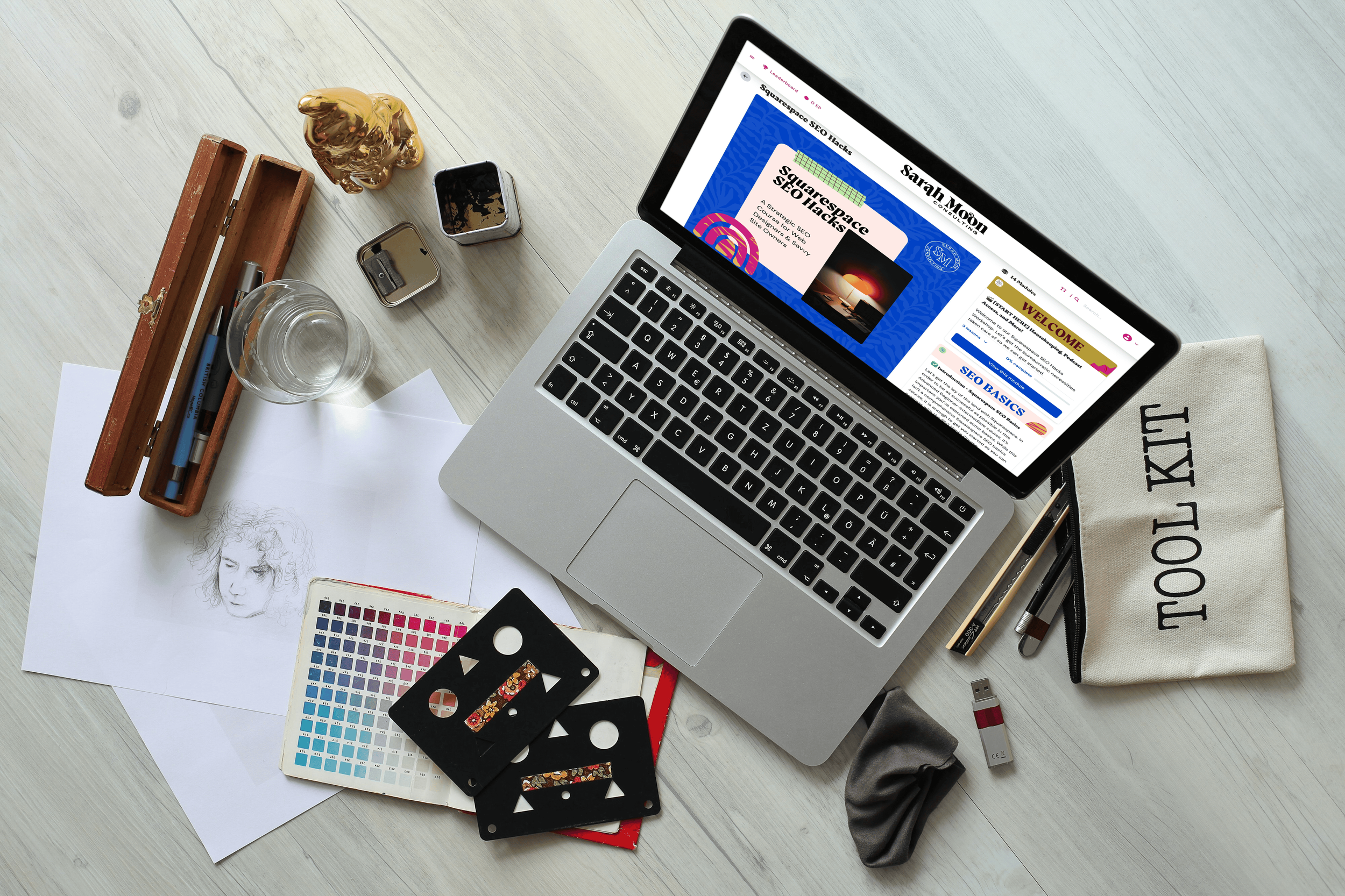 Overhead view of a workspace with a laptop open to a website about SEO, surrounded by drawing sketches, color palettes, pens, a glass of water, a USB flash drive, a scrunchie, and a cloth bag labeled 'tOOL KIT'.
