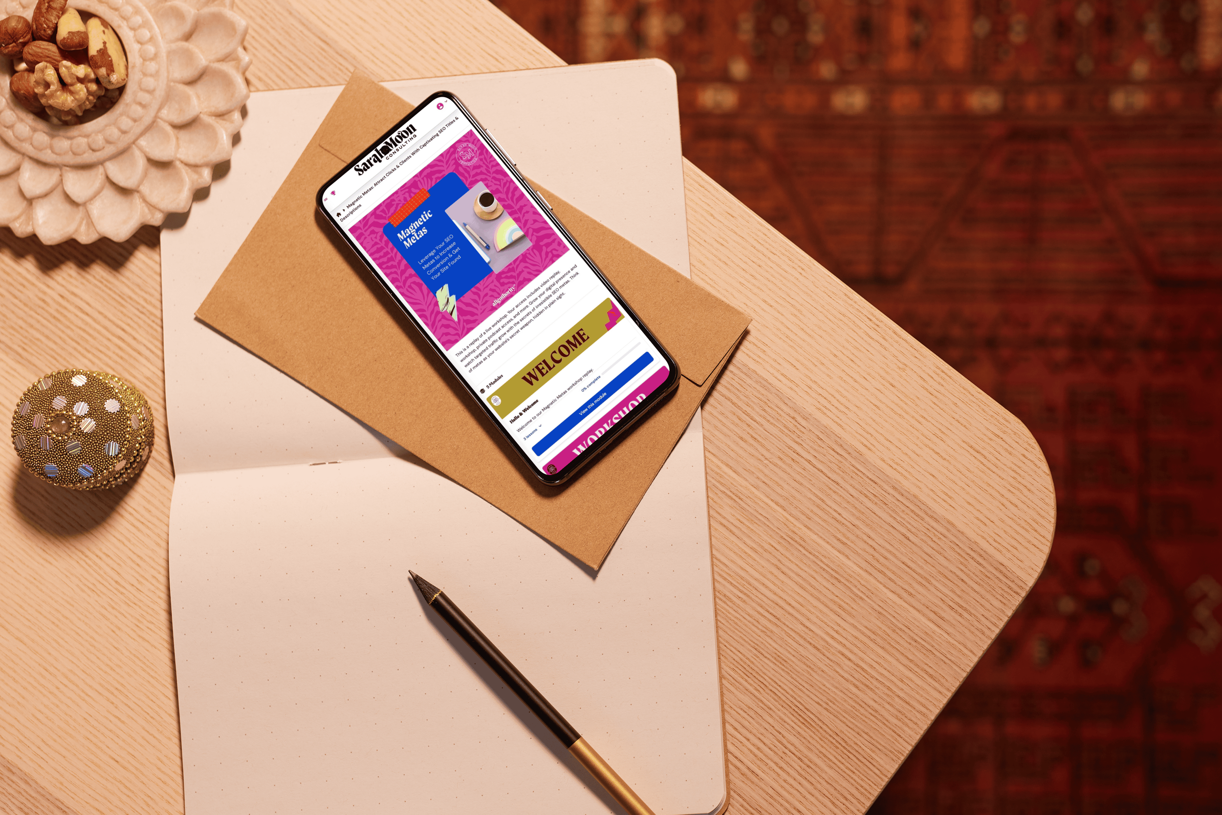 An iPhone placed on a brown envelope on a white and a beige paper on a wooden desk. The phone's screen shows a colorful webpage. Nearby are a black pen, a decorative gold and white ball, and a ceramic bowl with nuts. The background features a patterned red carpet.