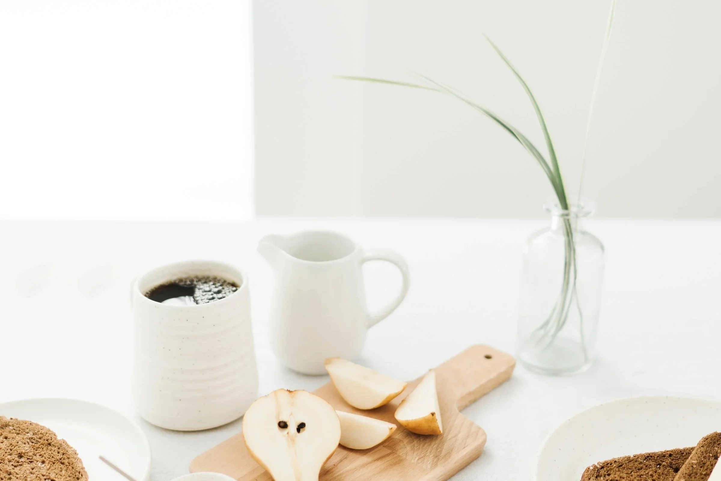 A minimalist breakfast setup with two white ceramic coffee cups, a small pitcher, sliced pears on a wooden board, and cookies on white plates, with a green plant in a clear glass vase in the background.