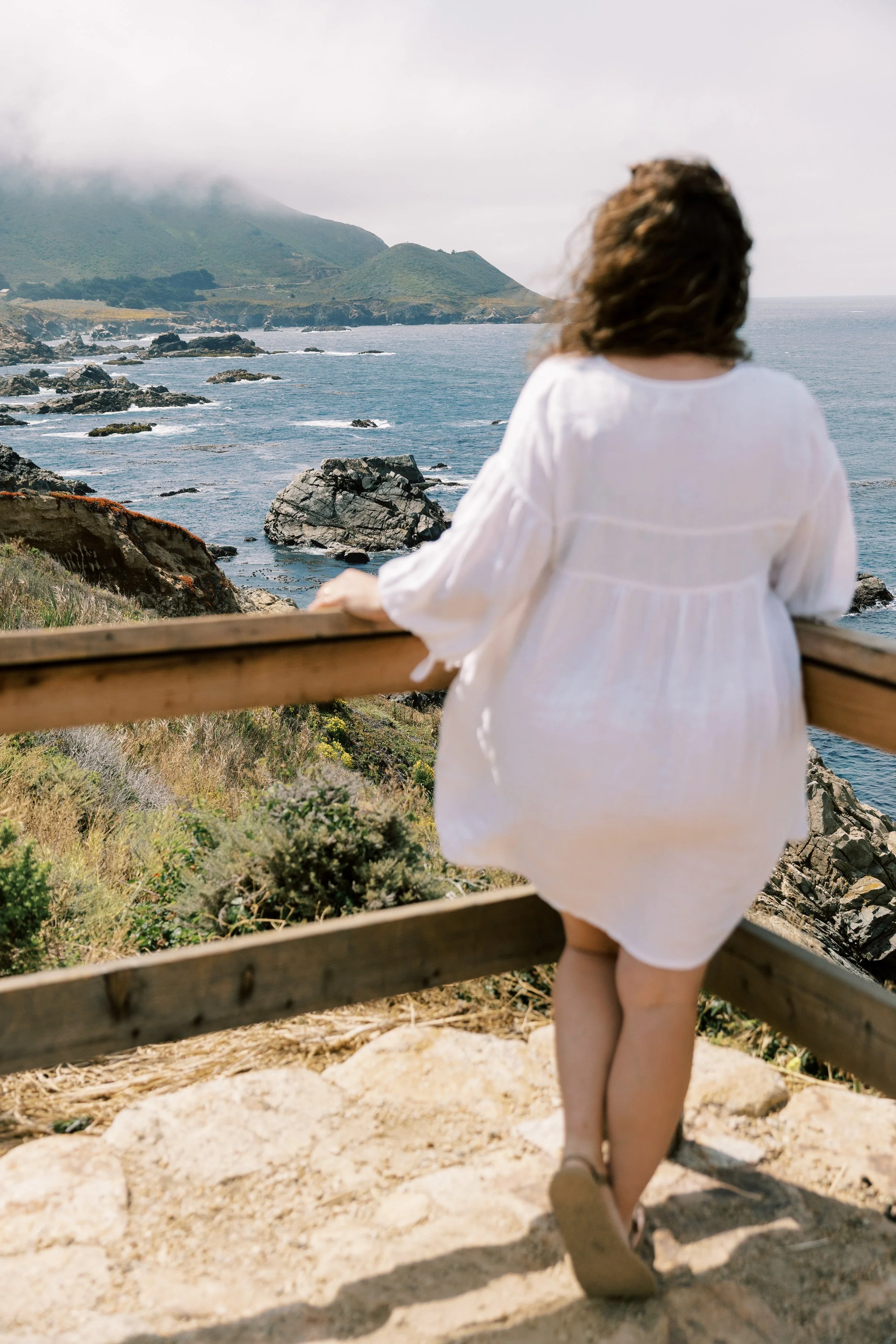A woman with long curly hair wearing a white dress stands on a wooden walkway overlooking a rocky coastline with mountains in the distance and an overcast sky.