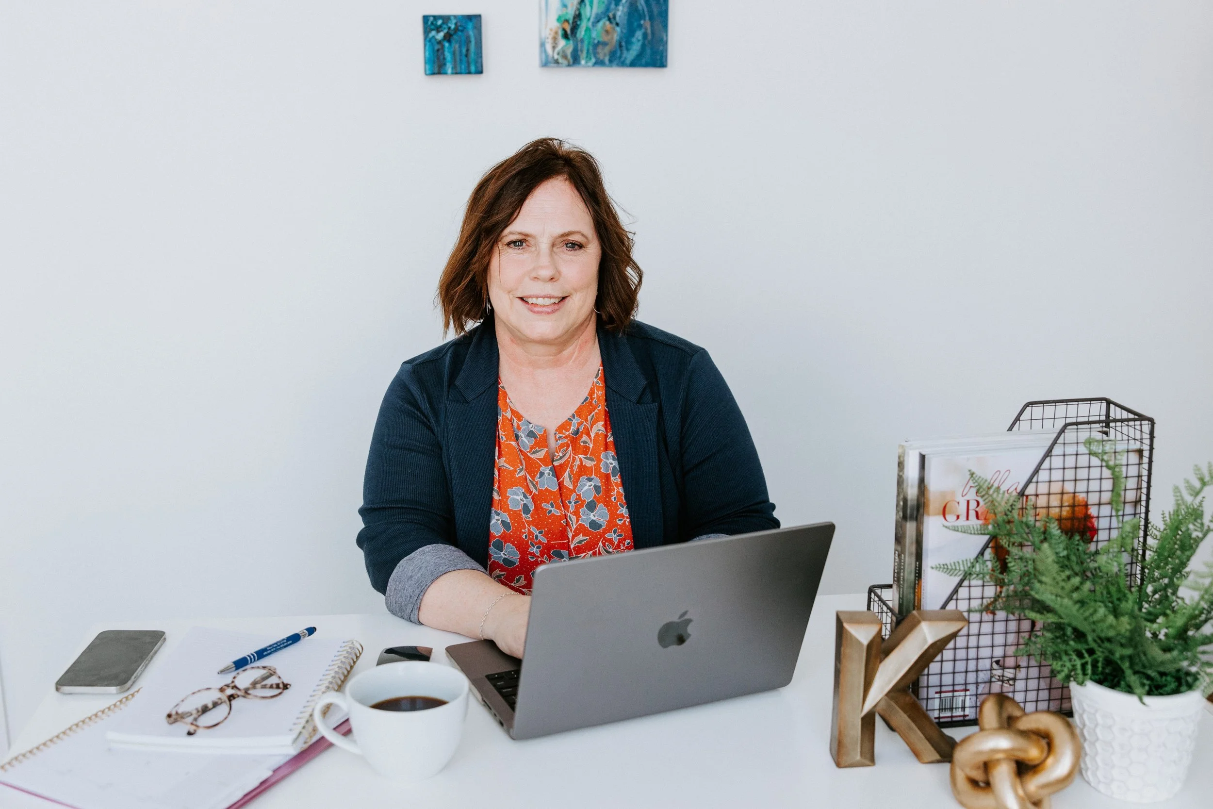 A woman at a white desk working on a silver laptop, surrounded by notebooks, a coffee mug, a smartphone, and stationery, with colorful abstract paintings on the wall behind her and a display of plants and decor items on her right side.