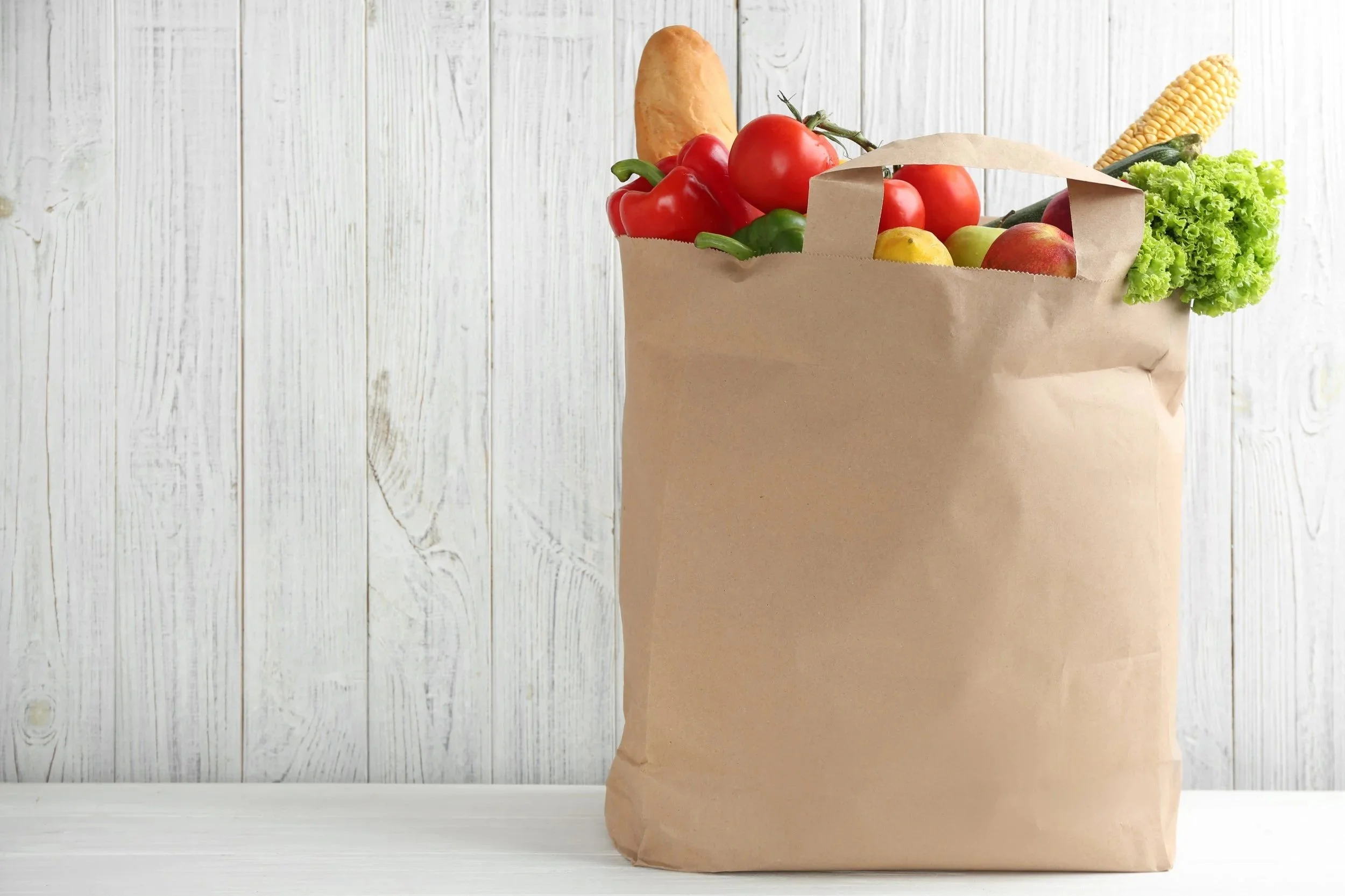 Paper grocery bag filled with fresh vegetables including lettuce, tomatoes, bell peppers, corn, zucchini, and a sweet potato, against a white wooden background.