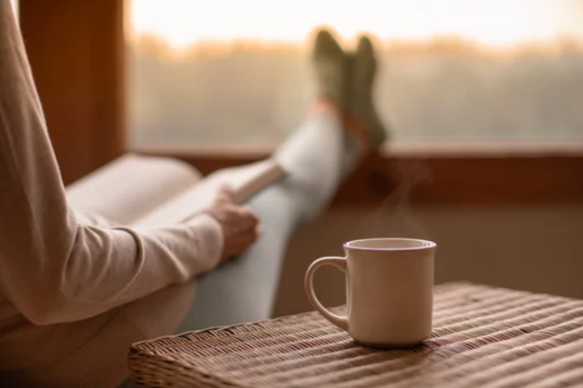 A person relaxing on a couch or bed with their legs stretched out, looking out a large window at sunset, holding a book. There is a white mug placed on a woven table or cloth in the foreground.