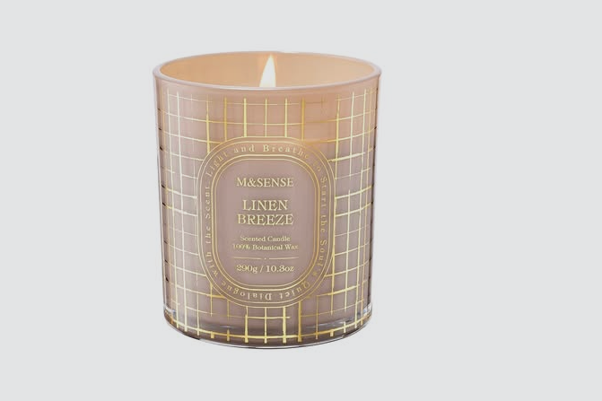 A lit scented candle in a decorative glass holder with a grid pattern and gold accents, labeled 'Linen Breeze' by M&SENSE.