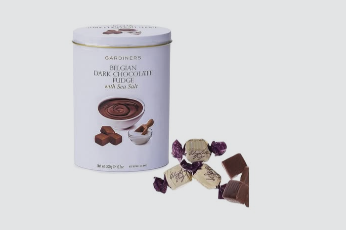 A cylindrical tin container of Gardiners Belgian Dark Chocolate Fudge with Sea Salt next to pieces of chocolate and wrapped candies.