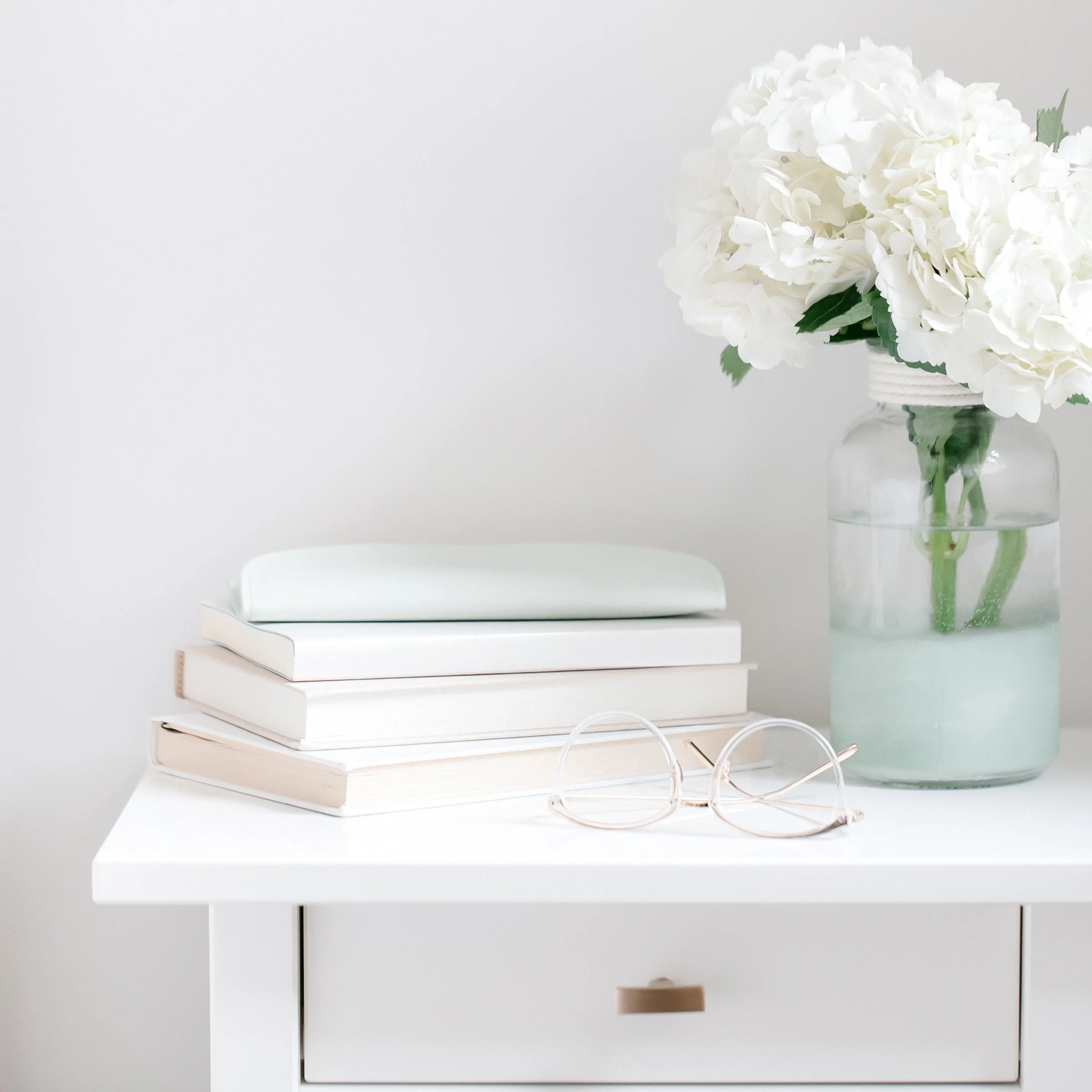 White hydrangea flowers in a glass vase on a white table against a light gray wall, with a stack of white books and a pair of pink eyeglasses nearby.