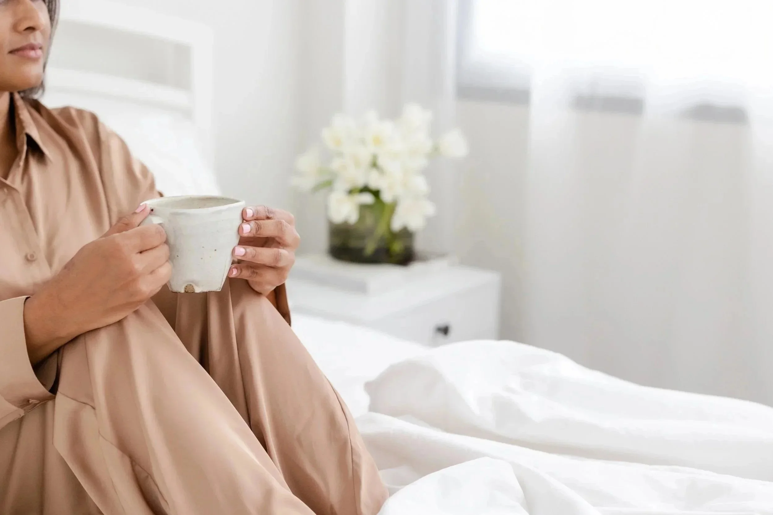 A woman in a beige dress sitting on a bed, holding a white ceramic mug, with a blurred background featuring a window and a flower vase.
