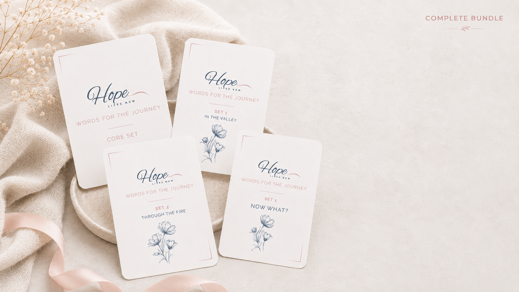 A set of four white cards with blue and pink text and floral illustrations, arranged on a beige ceramic dish and a soft, textured cloth, with a pink satin ribbon nearby. The cards are part of a bundle themed "Hope Lives Now" with sections titled "Words for the Journey" and numbered sets with different phrases.