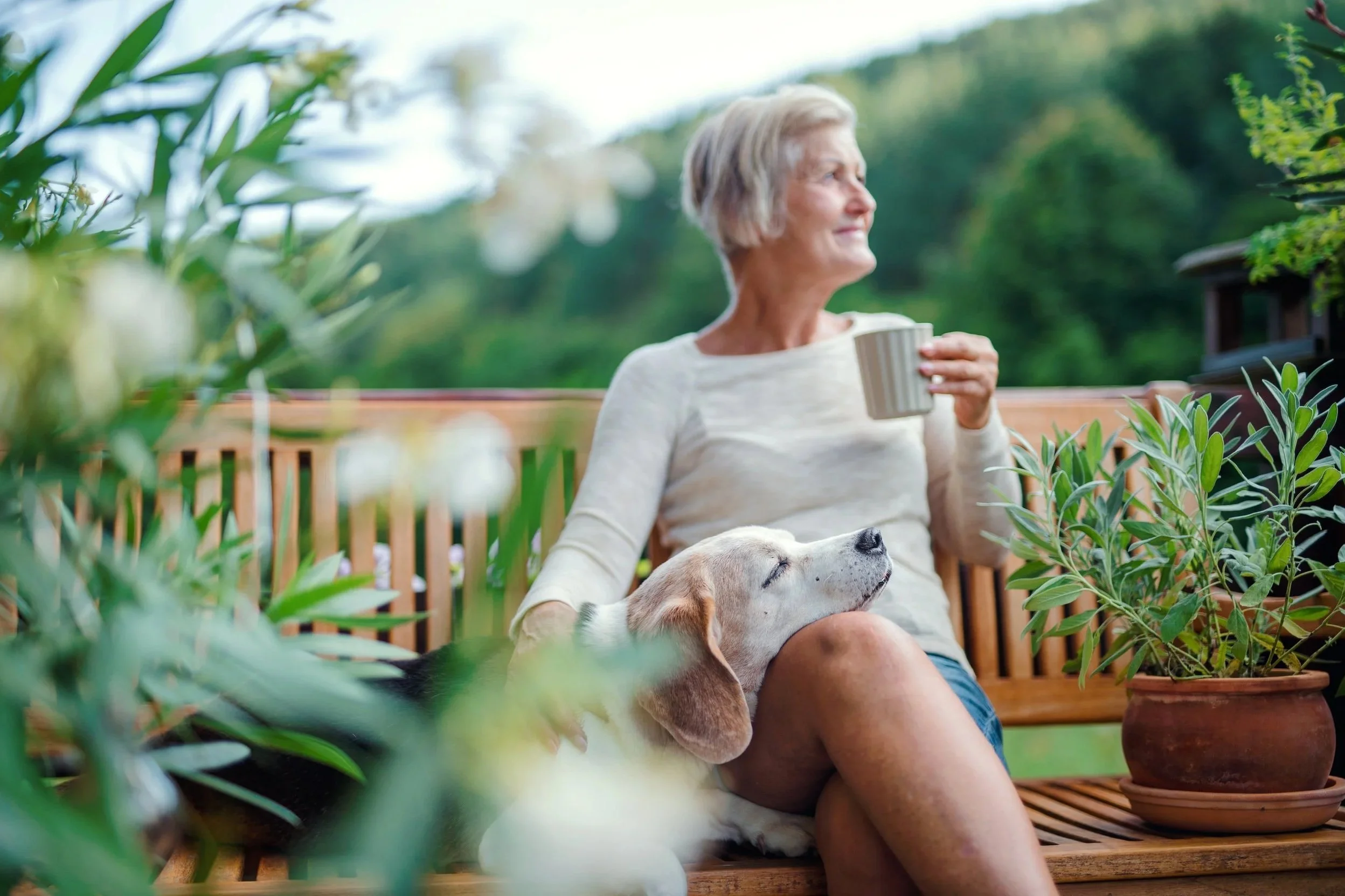 An older woman with gray hair sitting on a wooden outdoor bench, holding a cup of coffee and a beagle dog resting on her lap. She is surrounded by green plants in pots on a wooden deck in a lush garden setting.
