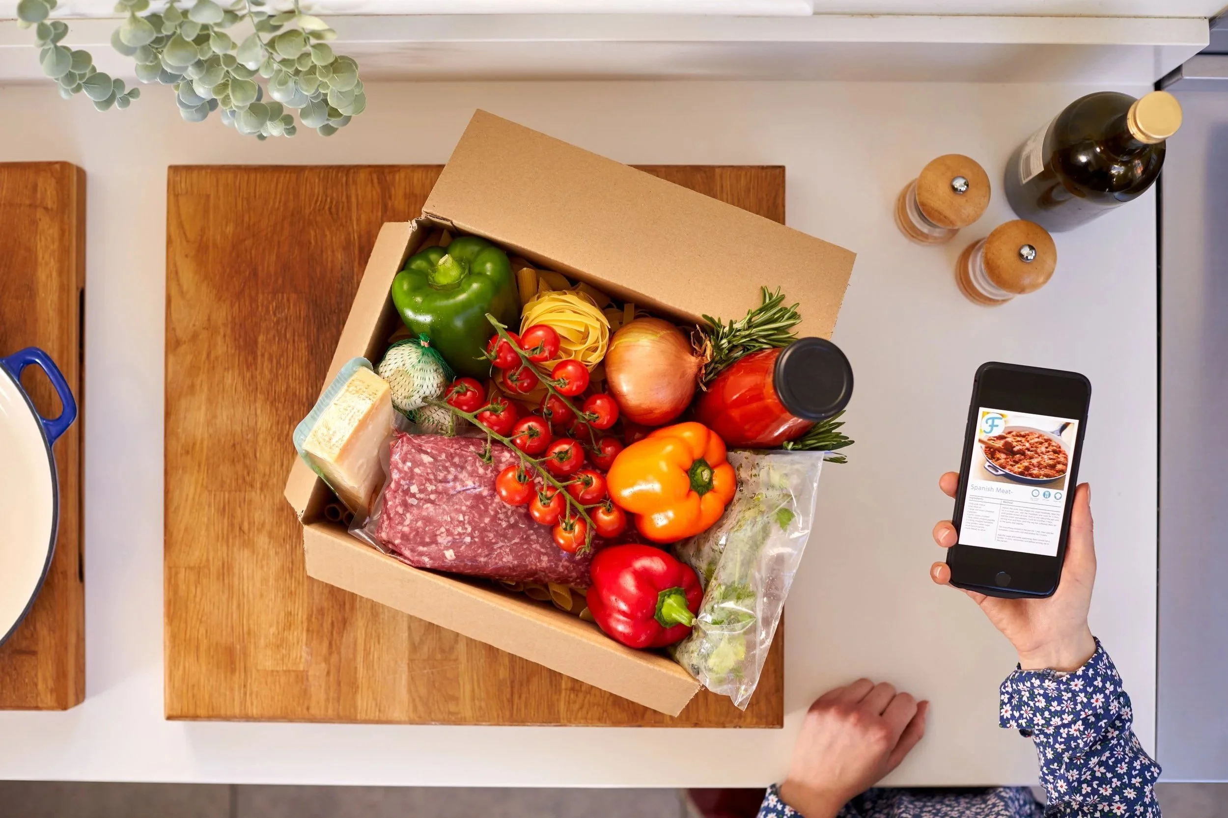A top-down view of a person taking a photo of a box of fresh groceries on a white table. The groceries include a green bell pepper, cherry tomatoes, a yellow bell pepper, an onion, a head of garlic, a package of meat, a bunch of rosemary, and a sheet of pasta. The person holds a smartphone displaying a food recipe app. A potted plant is visible at the top left corner.