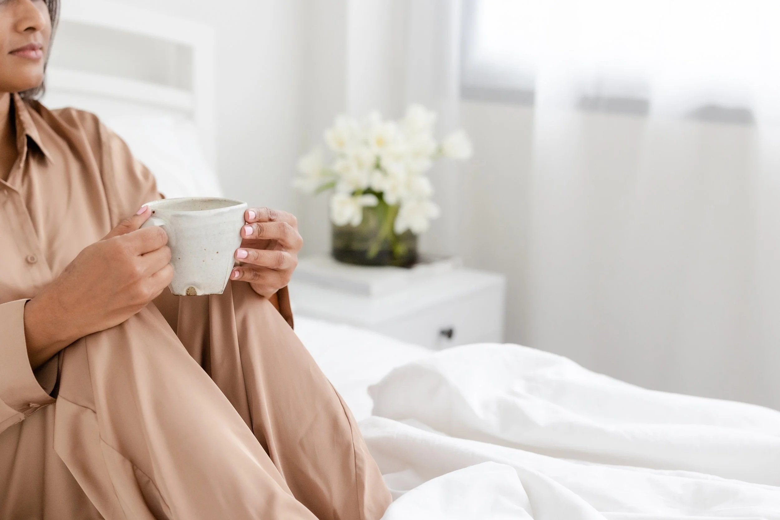 A person sitting on a bed holding a ceramic mug with both hands, wearing a beige satin shirt, in a bright, minimally decorated bedroom with a white flower arrangement on a side table.