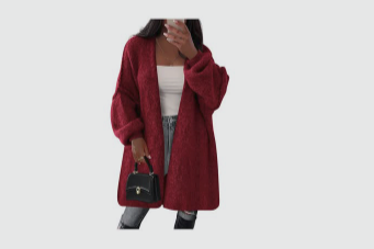 Person wearing a red cardigan, holding a black handbag, taking a mirror selfie with a smartphone, standing against a plain white background.