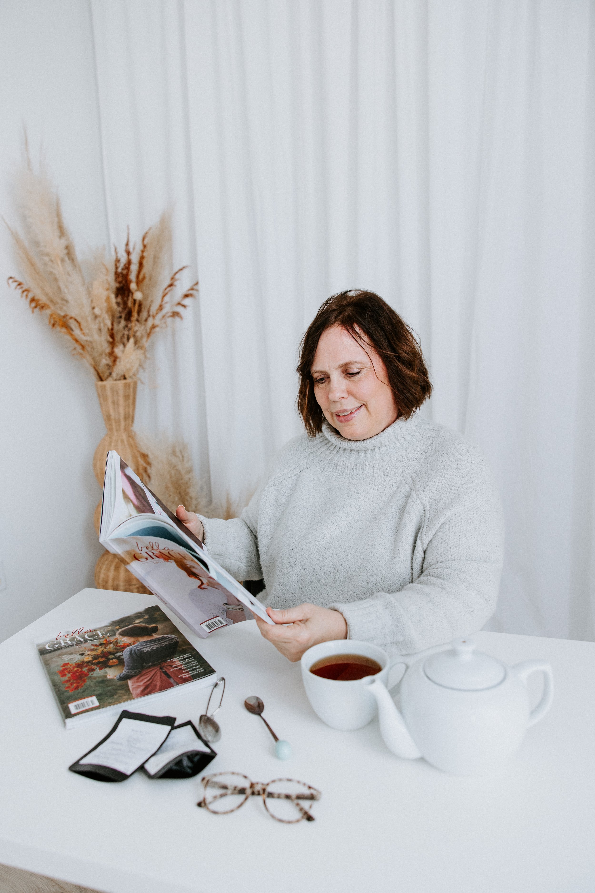 A woman in a gray sweater reads a magazine at a white table with various items, including a cup of tea, a book titled "Bella Grace," sunglasses, a beaded bracelet, and a notepad. In the background, a vase with dried pampas grass is visible against white curtains.