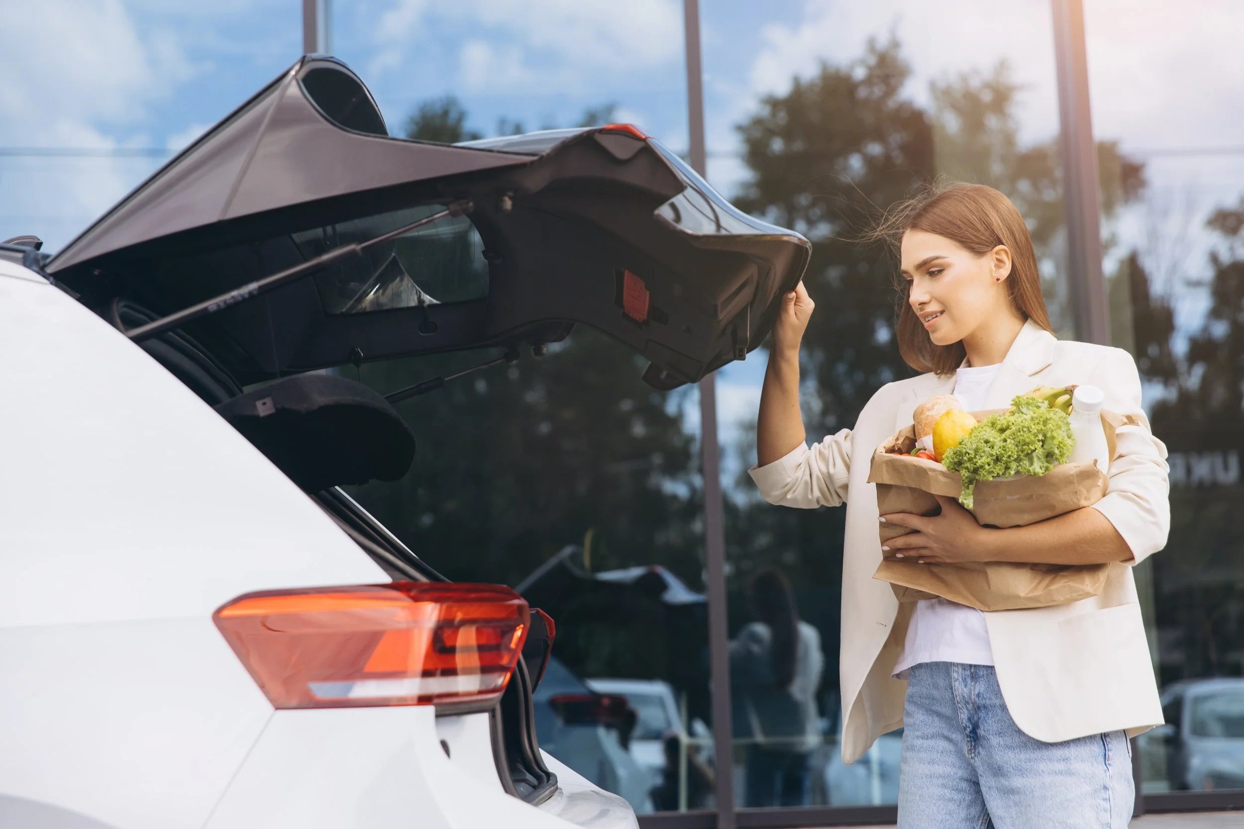 A woman with brown hair holding a grocery bag full of vegetables and groceries, standing by a white car with the trunk open, in a parking lot with trees and a cloudy sky in the background.